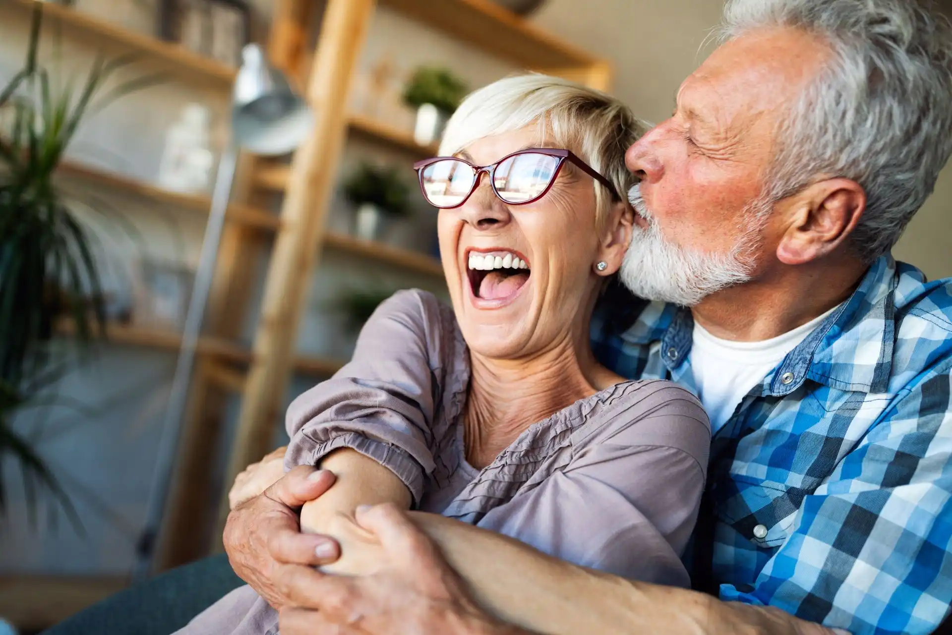 Smiling elderly man in blue plaid shirt hugging and kissing woman with short blonde hair and glasses.