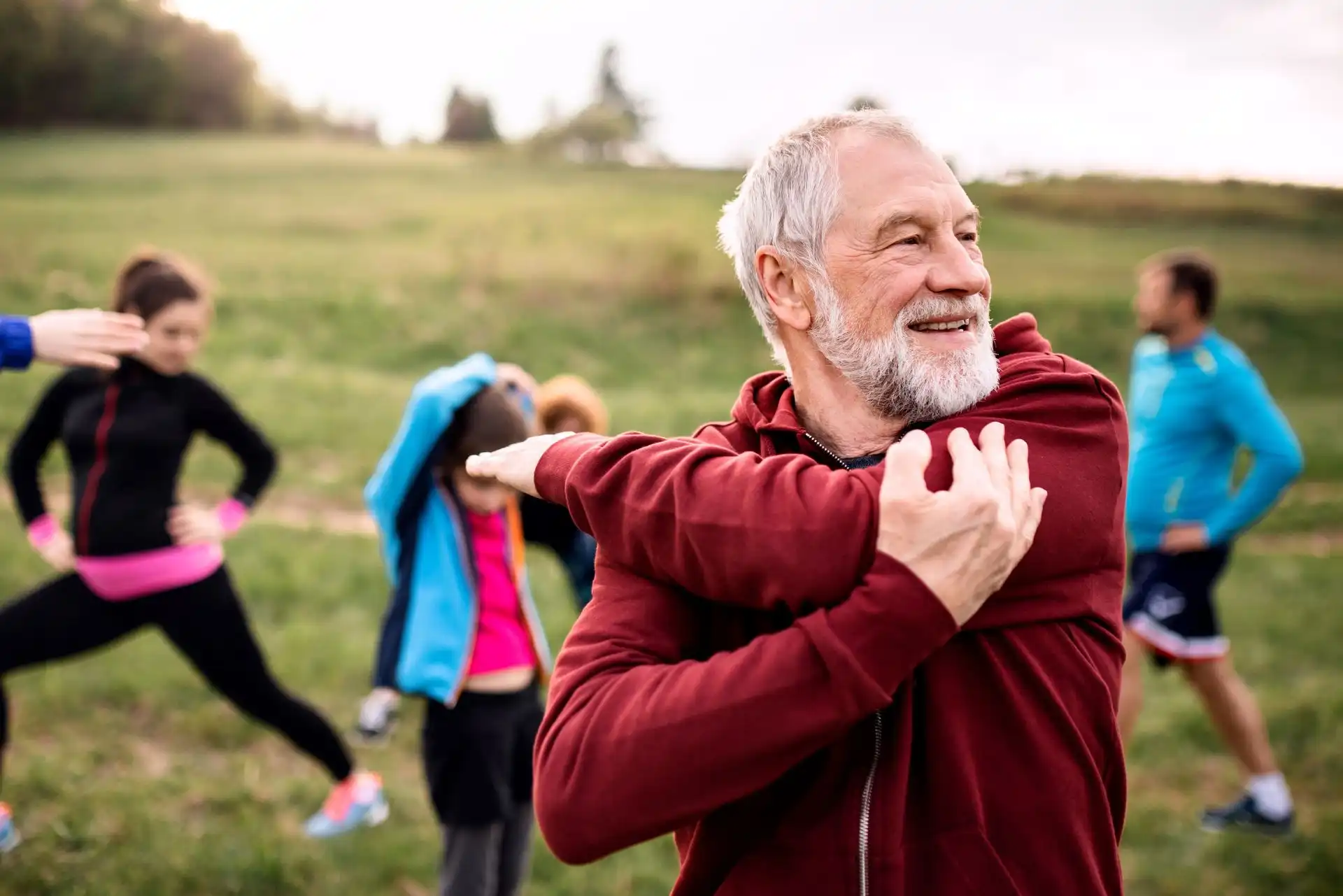 Smiling elderly man in a red jacket stretches his arm across his chest outdoors with a group exercising in the background.