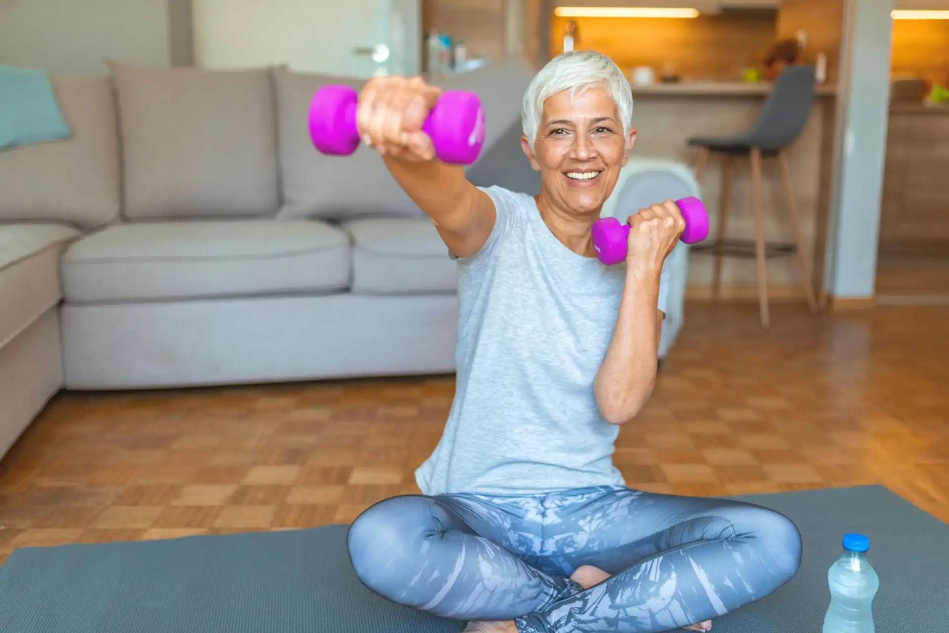 Smiling elderly woman sitting cross-legged on a mat, exercising with purple dumbbells at home.