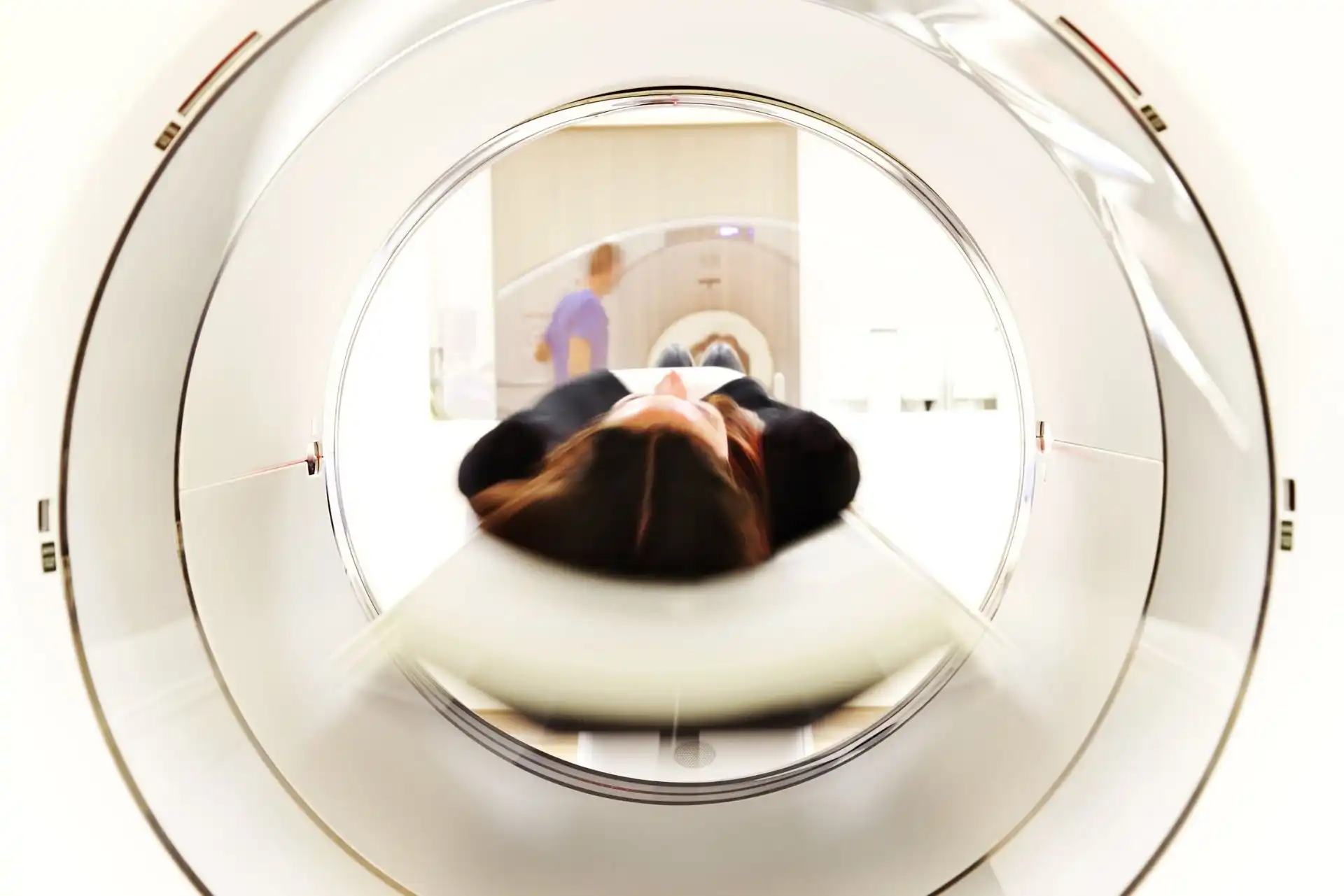 Patient lying on a sliding table entering an MRI machine for a brain scan while a technician monitors in the background.