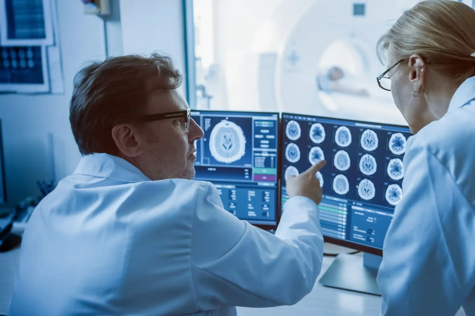 Two medical professionals in lab coats analyzing brain scan images on computer monitors in a diagnostic imaging room with an MRI machine in the background.