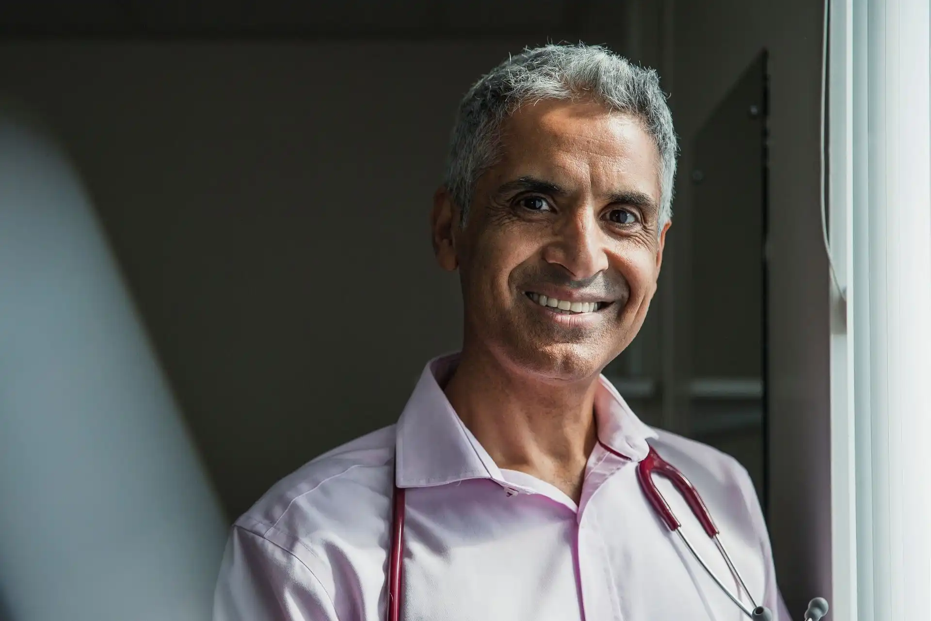 Smiling middle-aged male healthcare professional wearing a light pink shirt and stethoscope around his neck standing by a window.