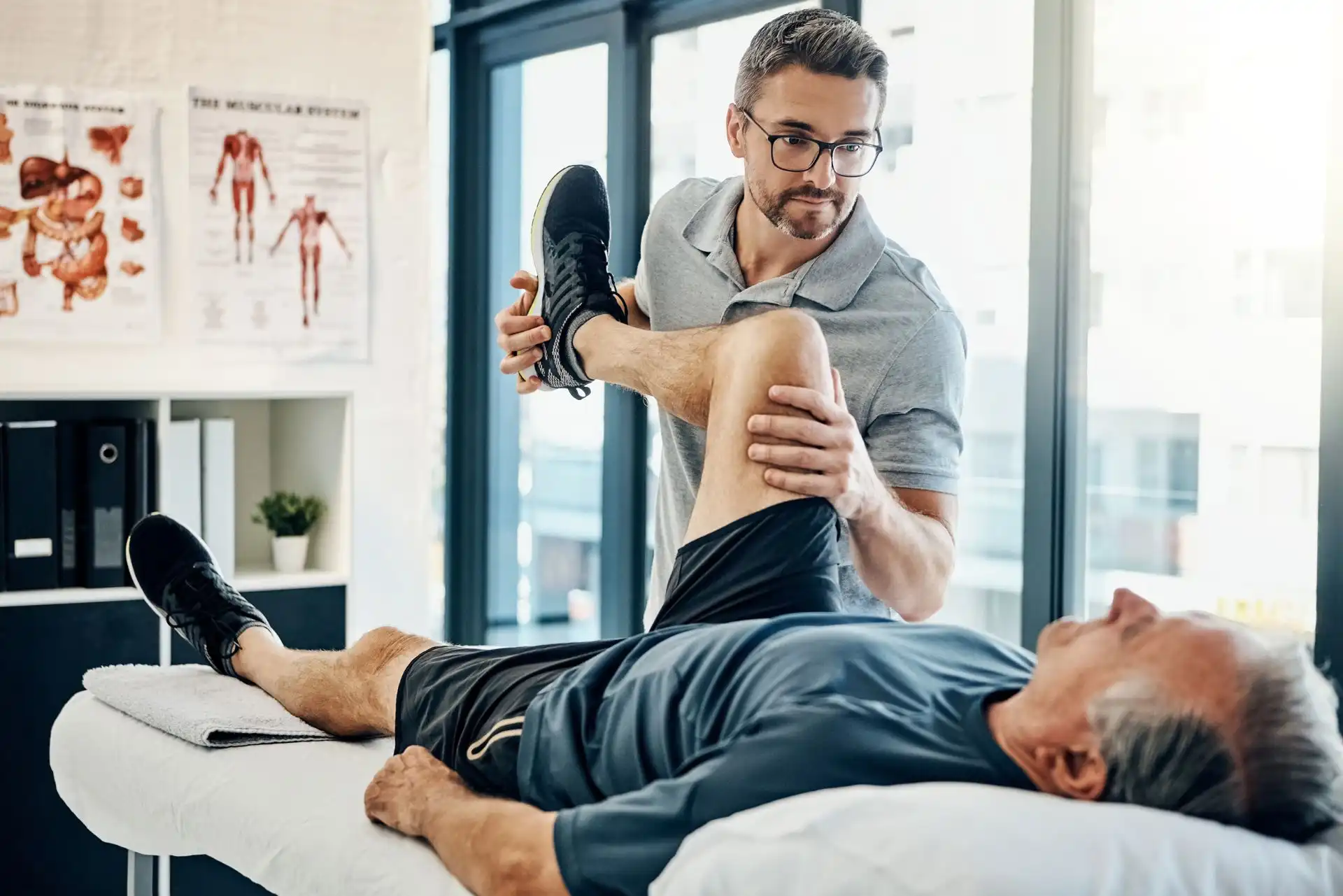 Physiotherapist assisting an elderly man with a leg stretch on a treatment table in a bright clinic.