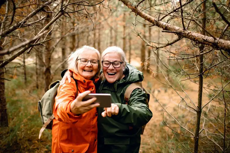 Two elderly women wearing outdoor jackets and backpacks smiling and taking a selfie in a forest during autumn.