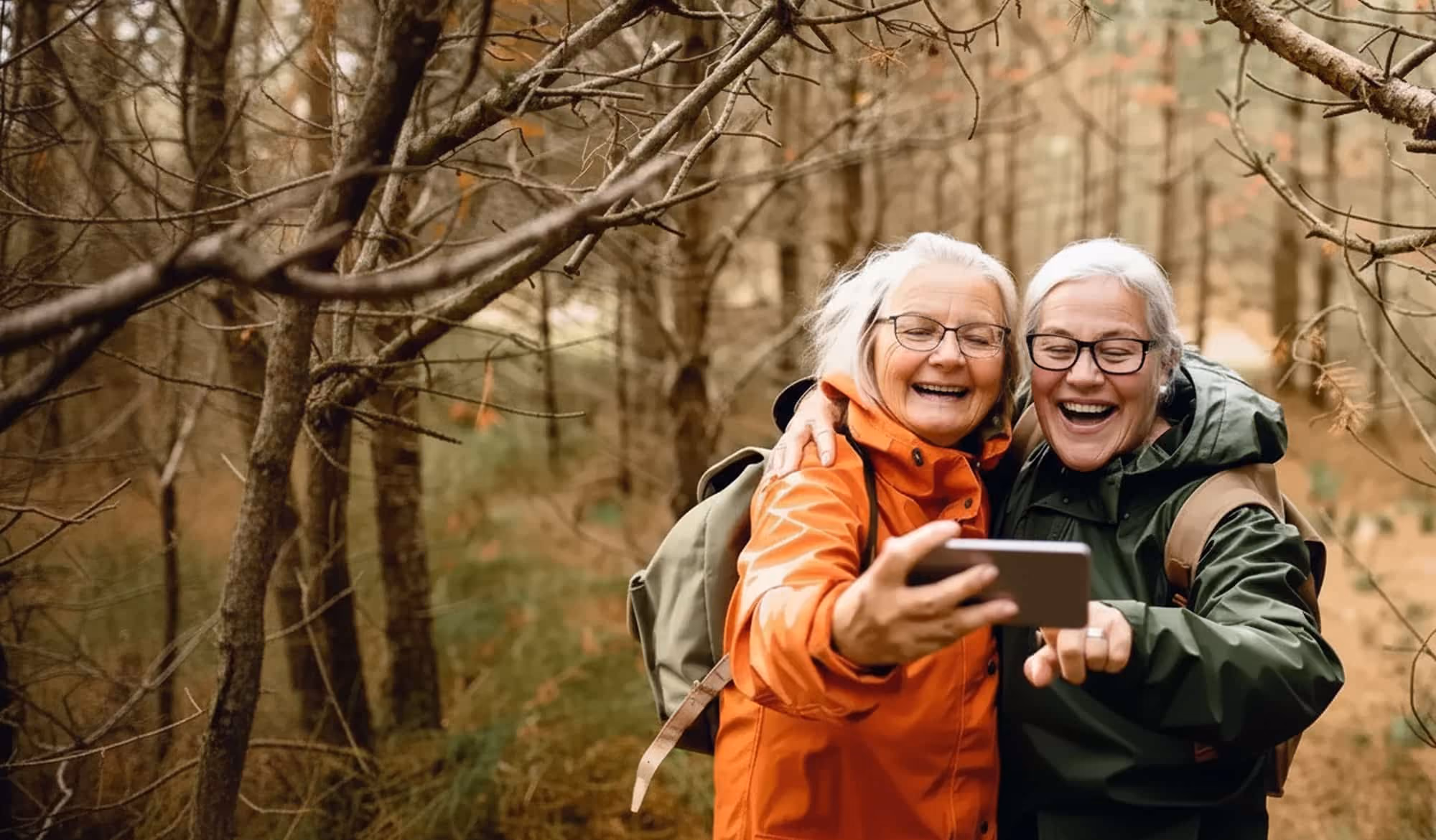 Two elderly women with gray hair and glasses taking a selfie together outdoors in a forest, wearing backpacks and jackets.