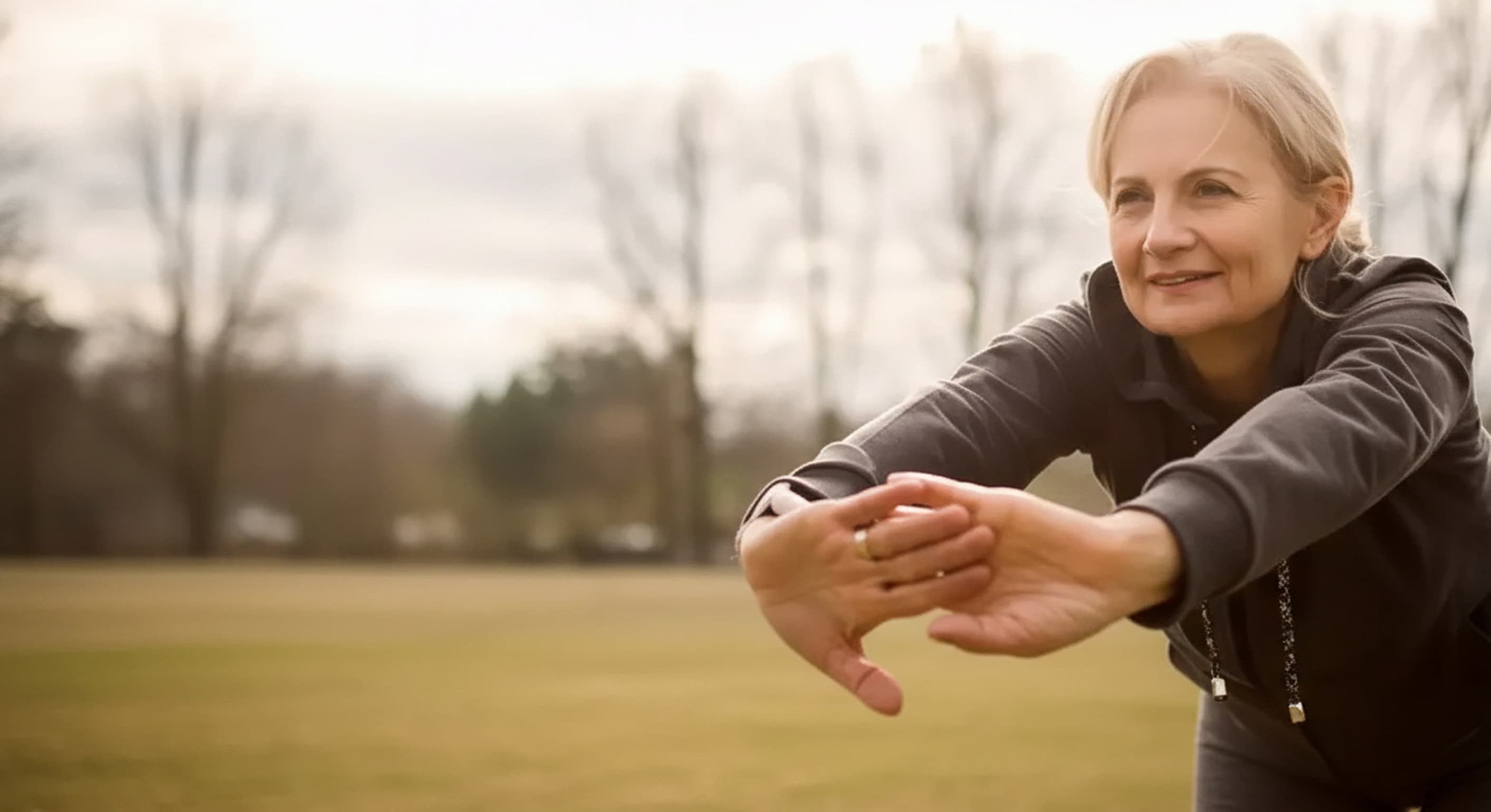 Smiling middle-aged woman stretching arms forward outdoors in a park.