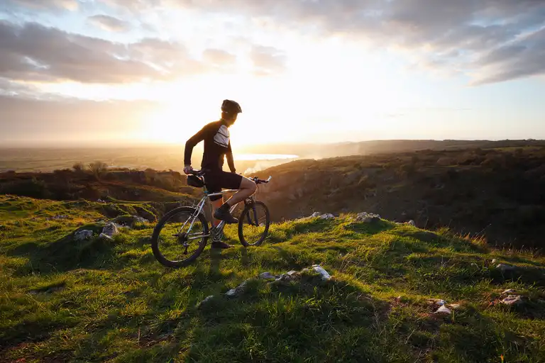 Cyclist wearing helmet standing with bike on a grassy hill at sunset overlooking a valley.