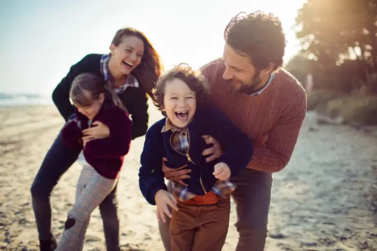 A smiling man and woman play and laugh with two young children on a sunny beach.