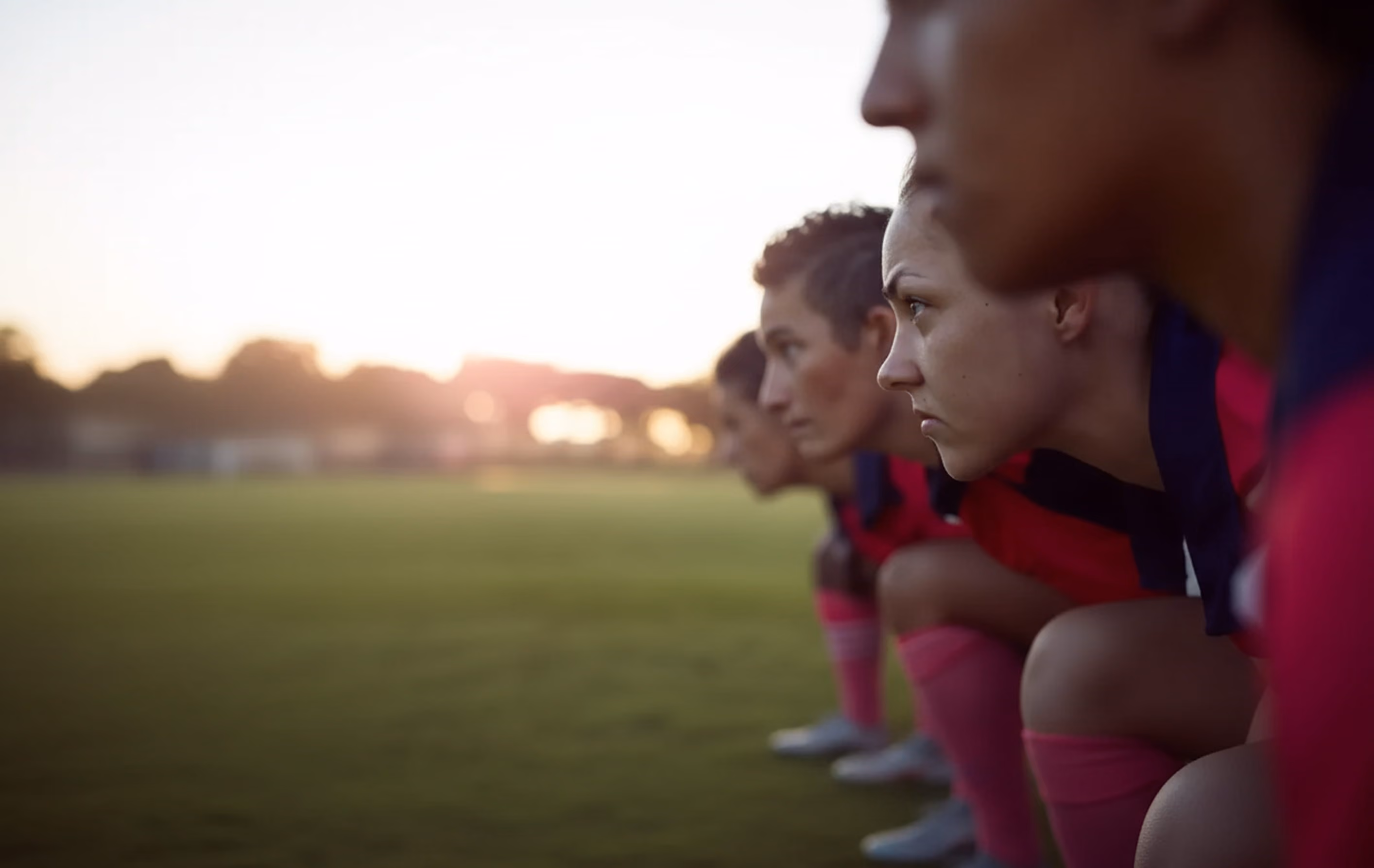 Close-up side view of female soccer players in a crouched starting position on a field at sunset.