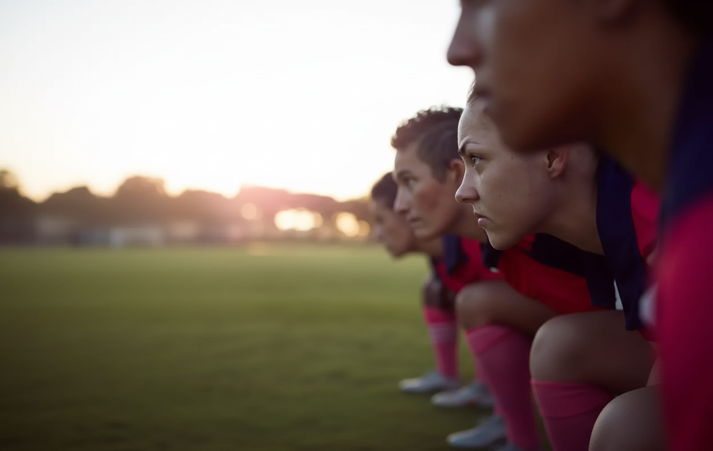 Close-up side view of female soccer players in a crouched starting position on a field at sunset.