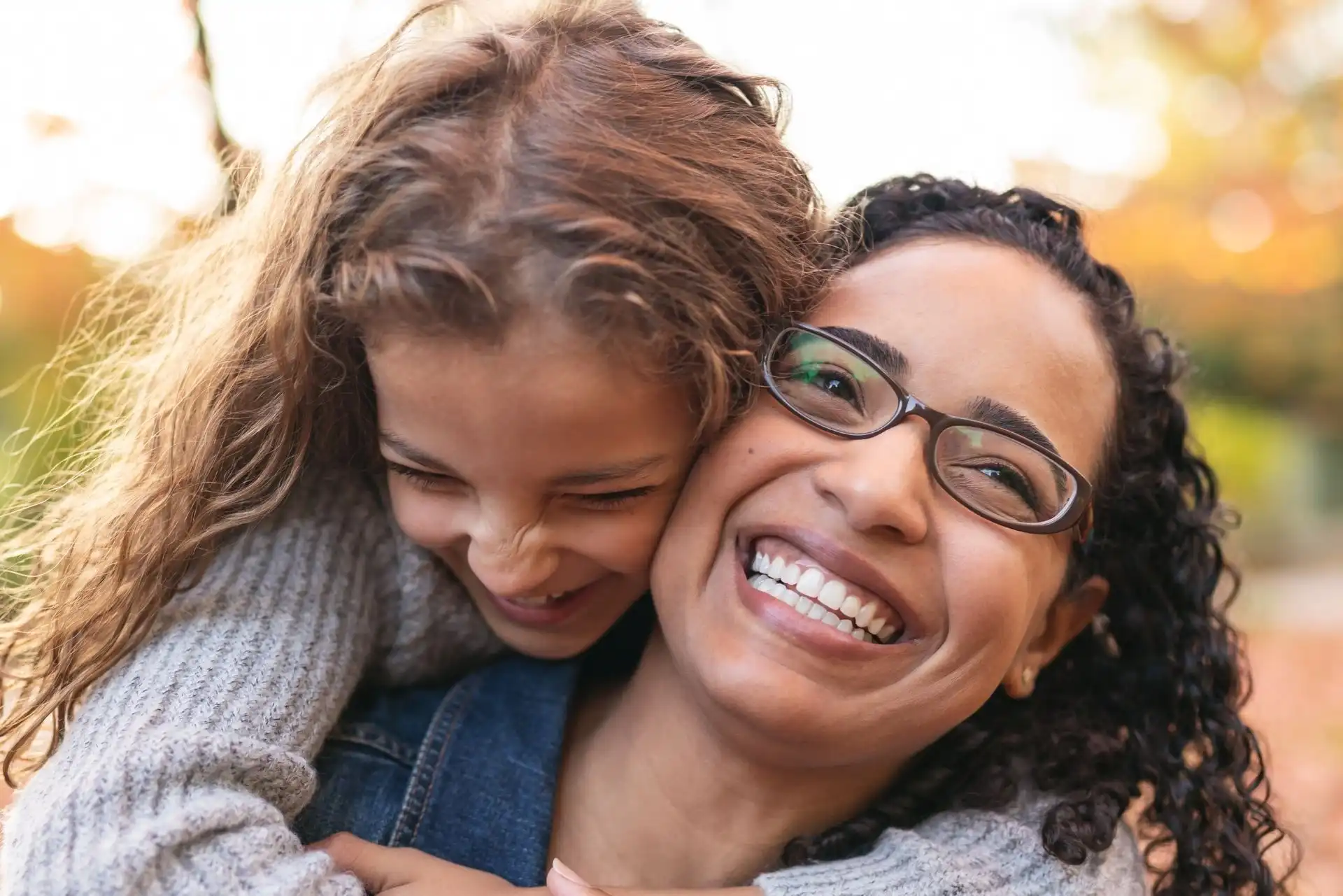 Smiling woman wearing glasses being hugged from behind by a laughing young girl with curly hair.