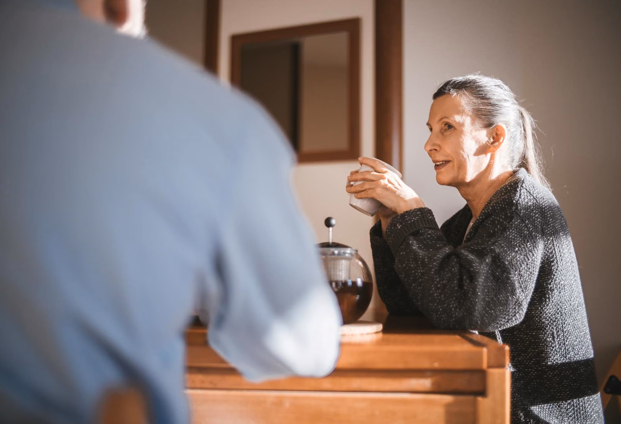 Older woman holding a cup and talking to a person sitting across a wooden table.