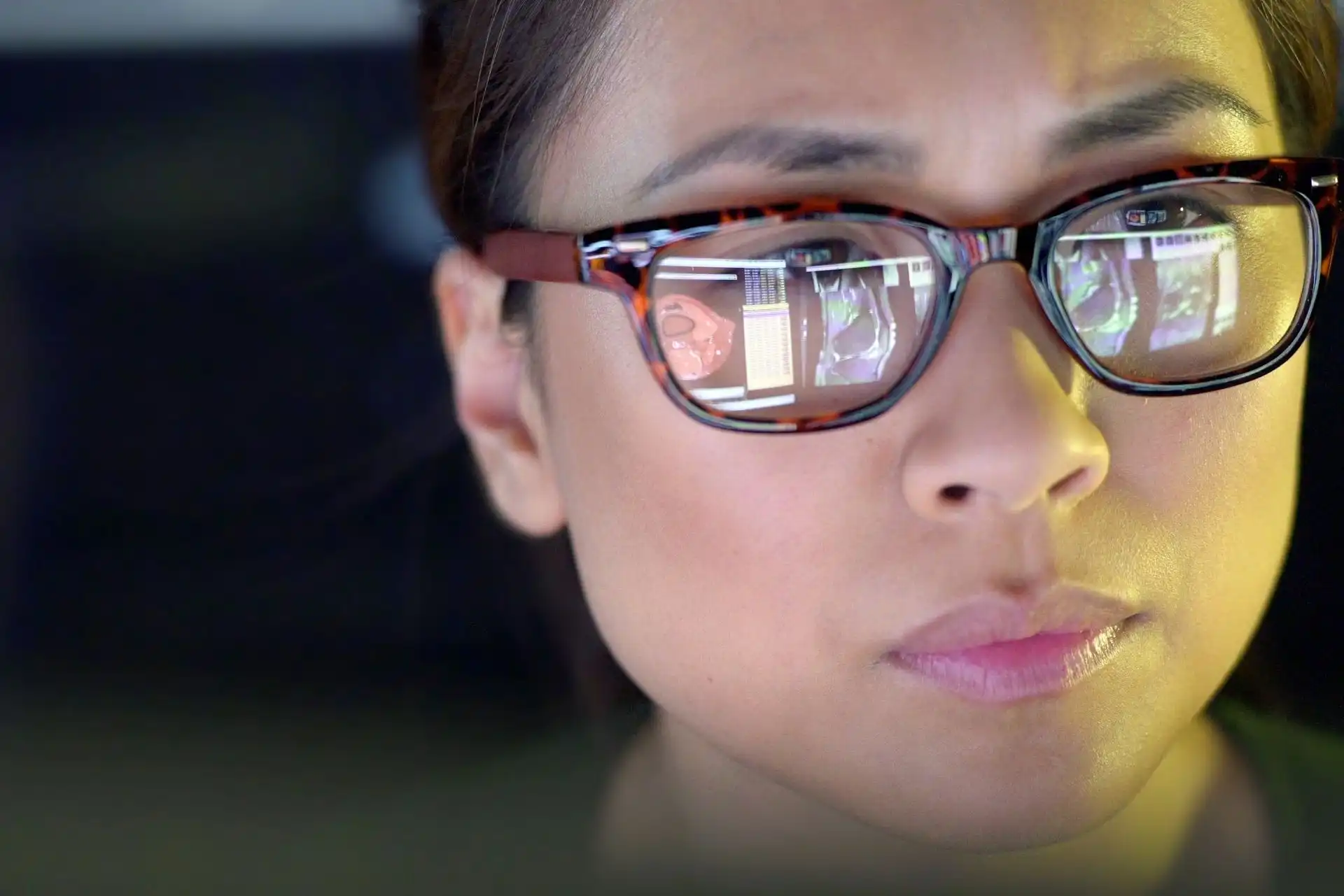 Close-up of a woman wearing tortoiseshell glasses reflecting medical imaging scans on a dark background.