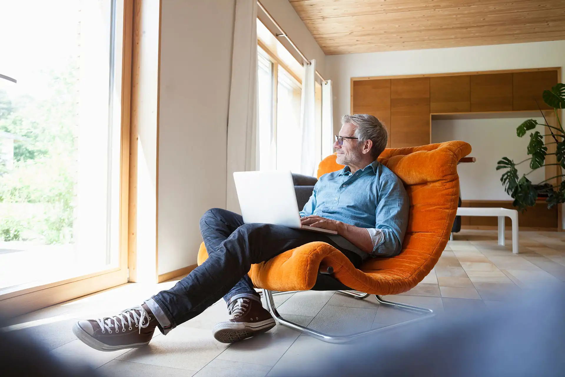 Middle-aged man in glasses sitting in an orange chair with a laptop, looking out a large window.