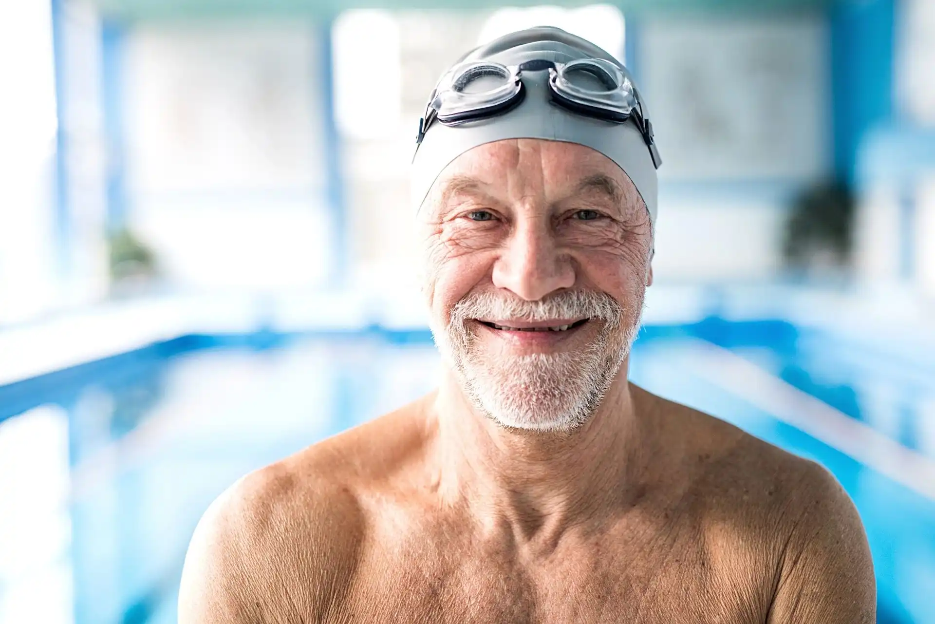 Smiling elderly man wearing a swim cap and goggles standing in an indoor swimming pool.