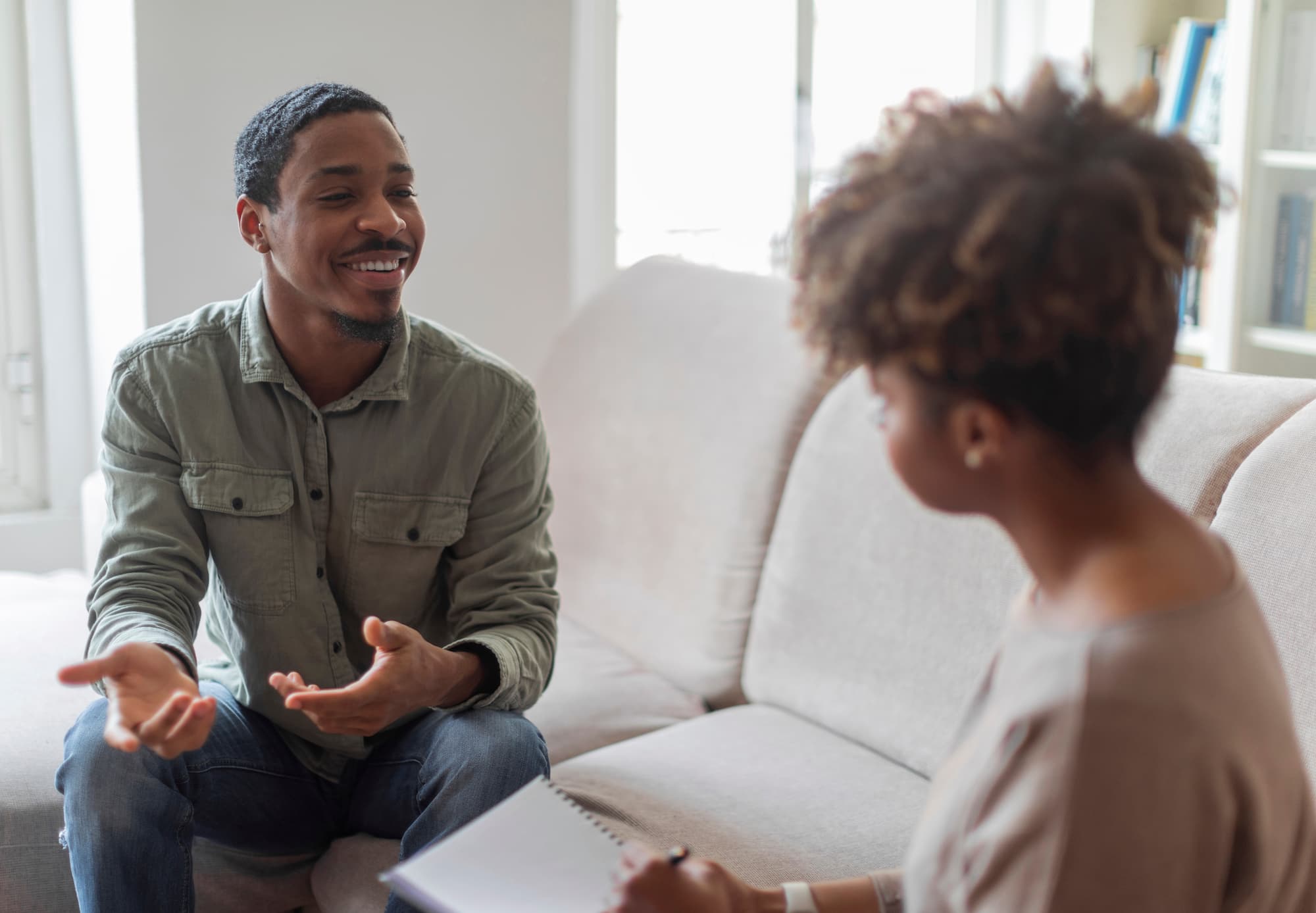Smiling man in casual clothes talking while seated on a sofa with a woman taking notes.