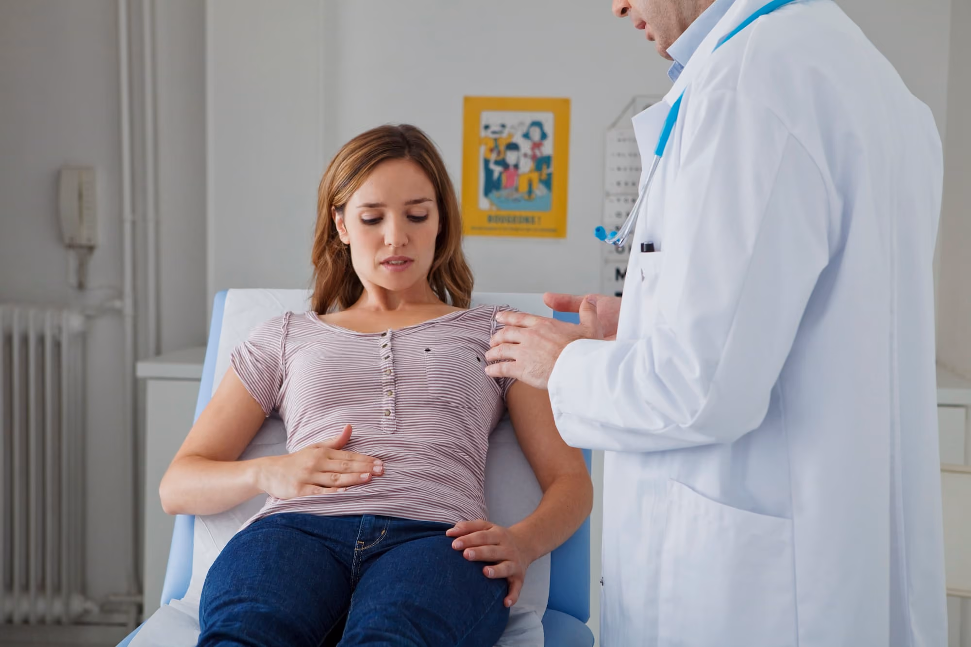 Young woman sitting on a medical examination table holding her abdomen while a doctor in a white coat speaks to her.
