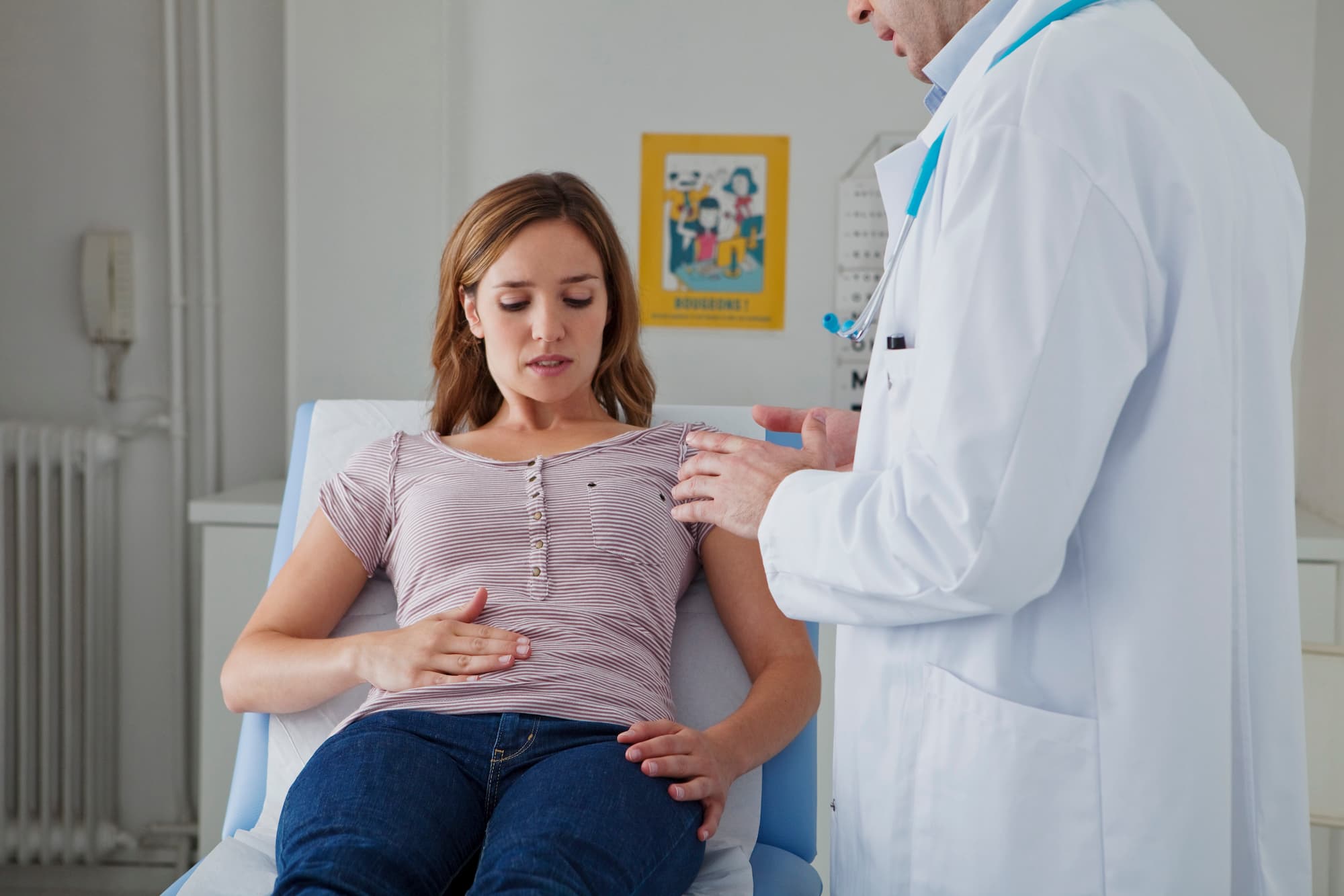 Young woman sitting on a medical examination table holding her abdomen while a doctor in a white coat speaks to her.
