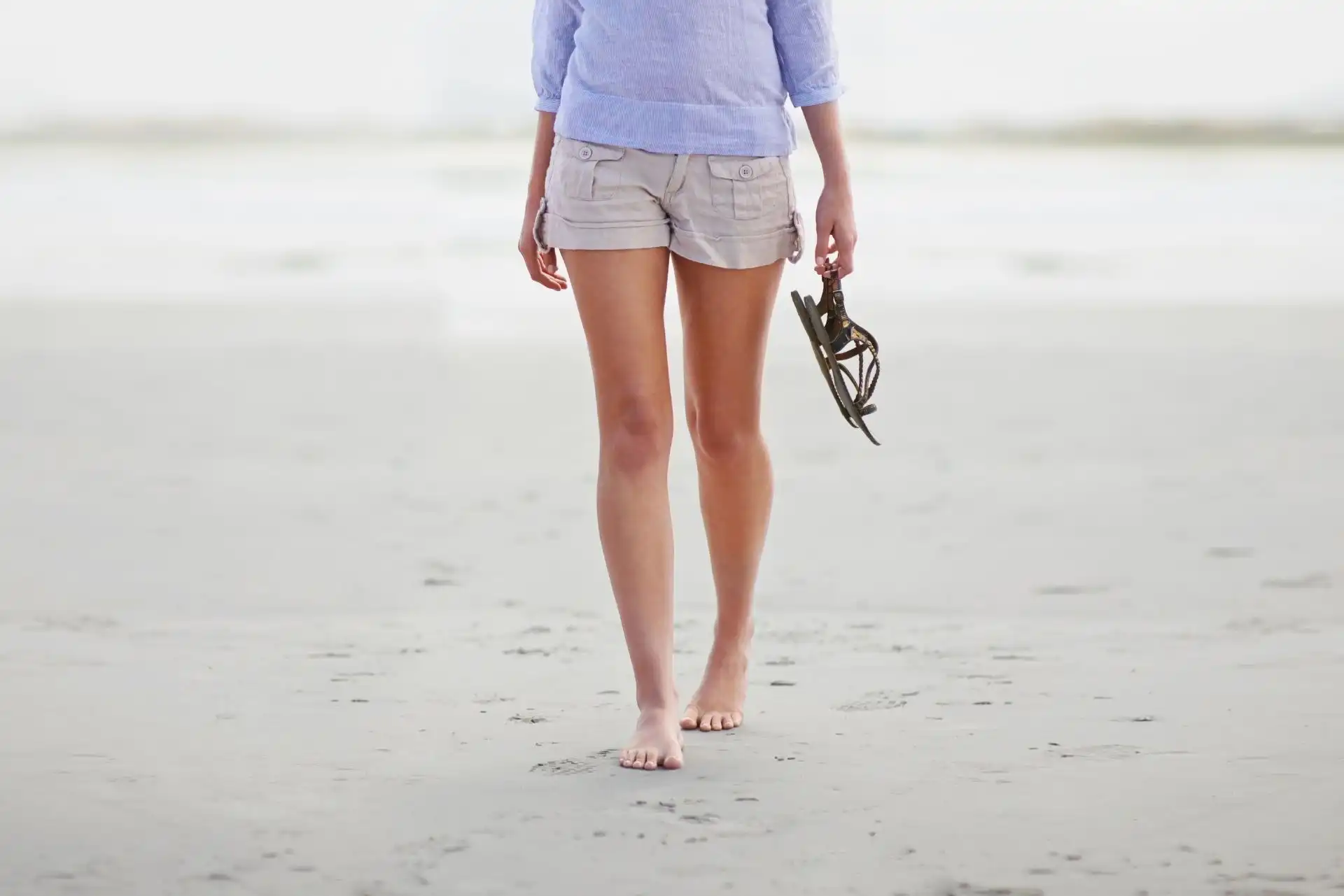 Person walking barefoot on a beach holding a pair of sandals in one hand.