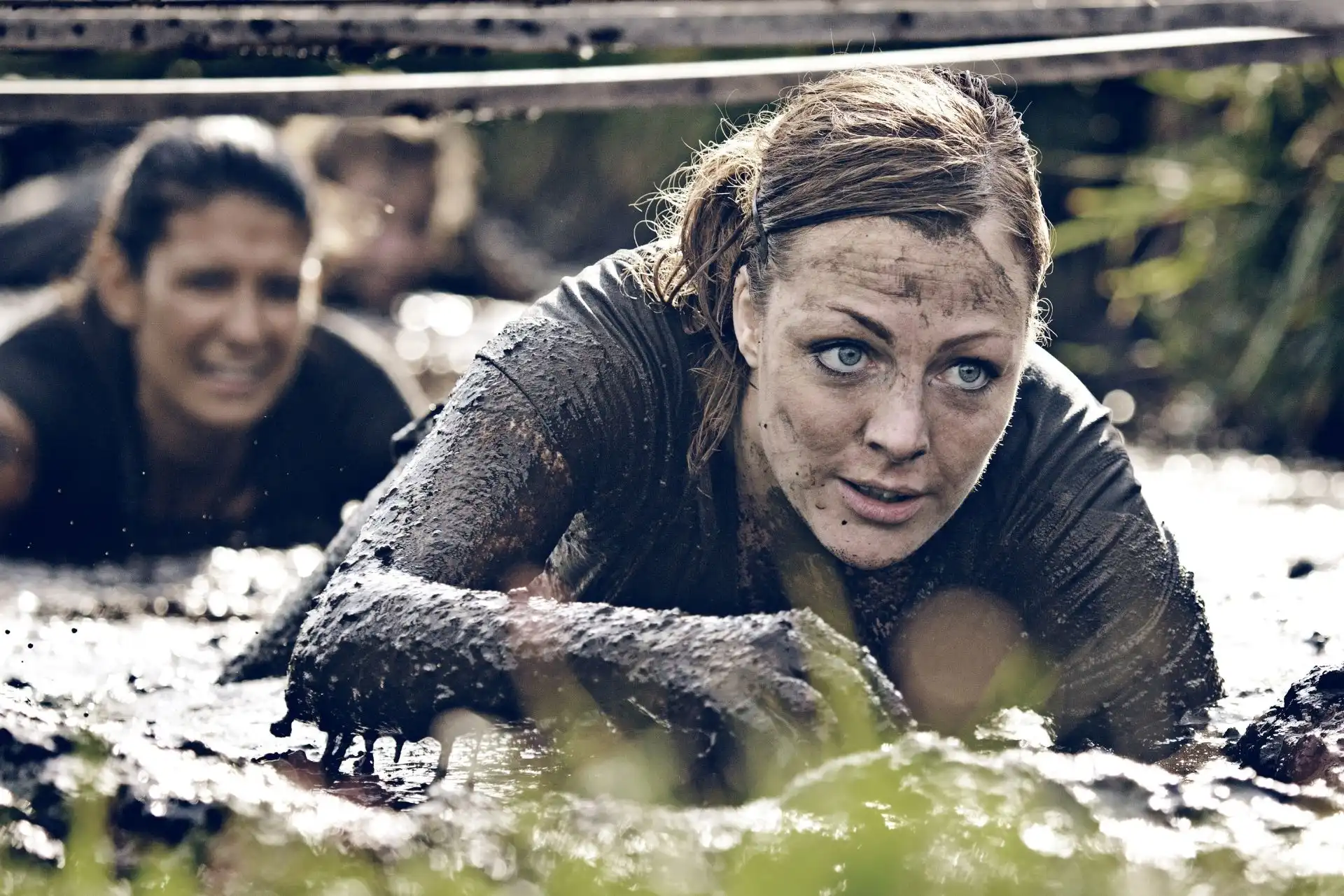 Woman covered in mud crawling through a muddy obstacle course outdoors with another person behind her.