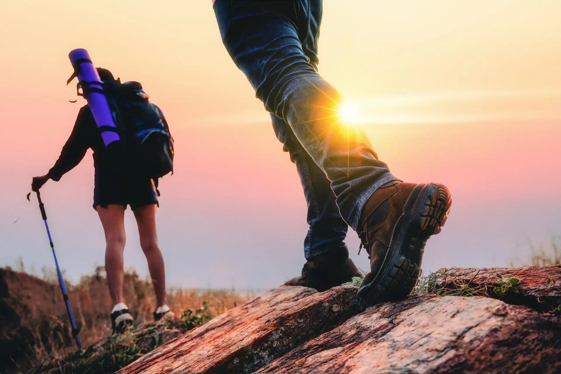 Two hikers on a rocky trail at sunset, one with a trekking pole and backpack, the other wearing brown hiking boots stepping on a rock with sunlight shining through.