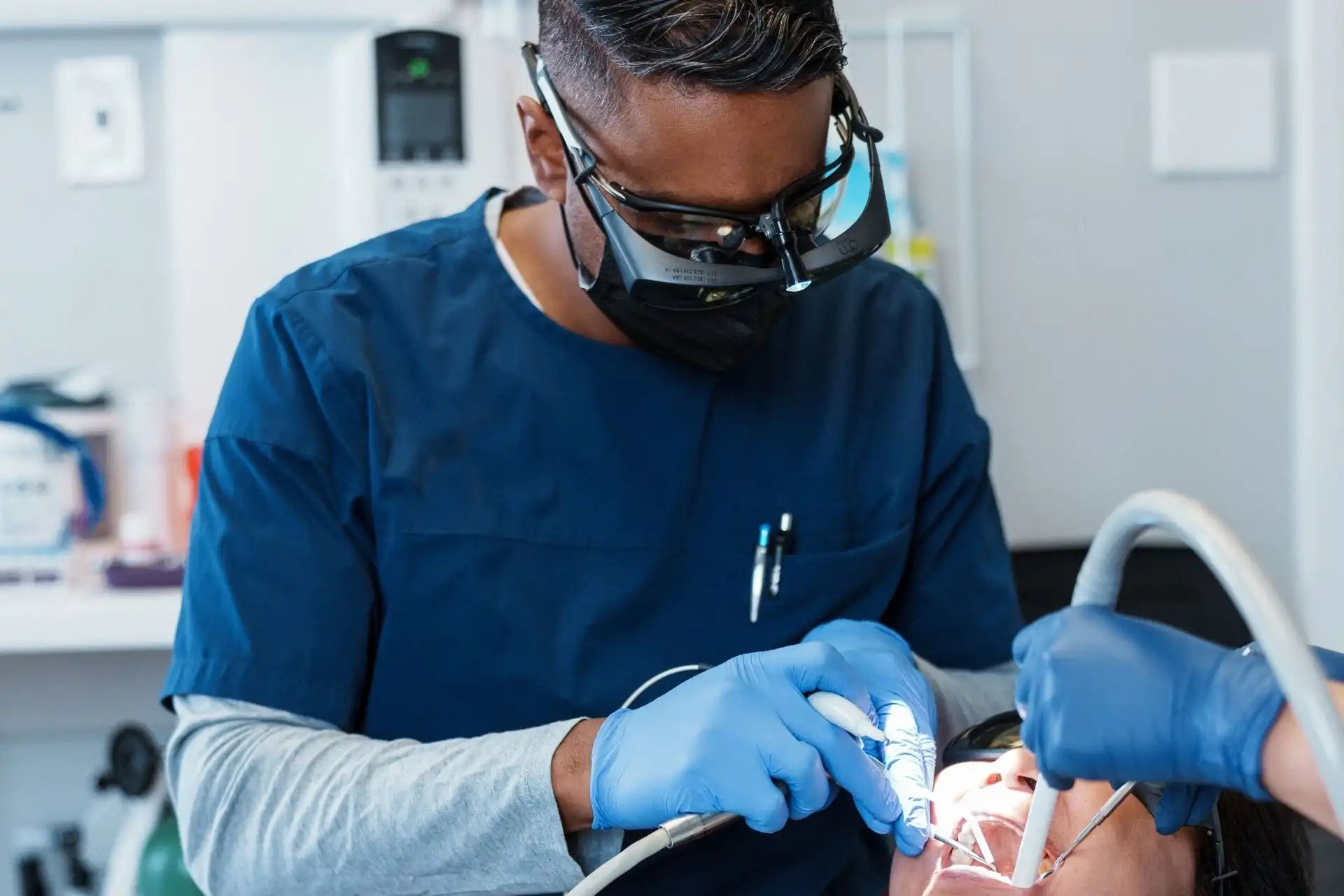 Dentist wearing magnifying glasses and a black mask performing a dental procedure on a patient.