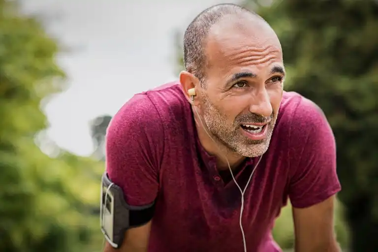 Middle-aged man in a maroon shirt with earbuds and armband leaning forward outdoors, looking determined.