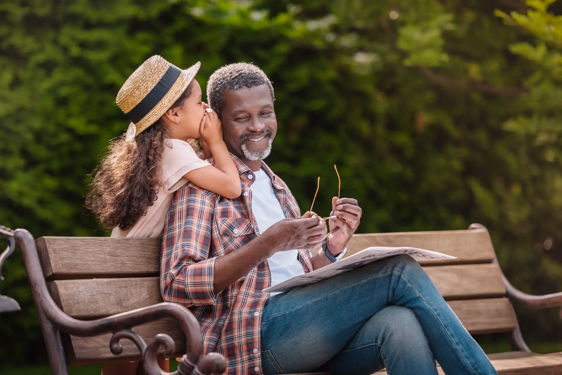Young girl whispering into her smiling father's ear while they sit on a wooden bench outdoors.