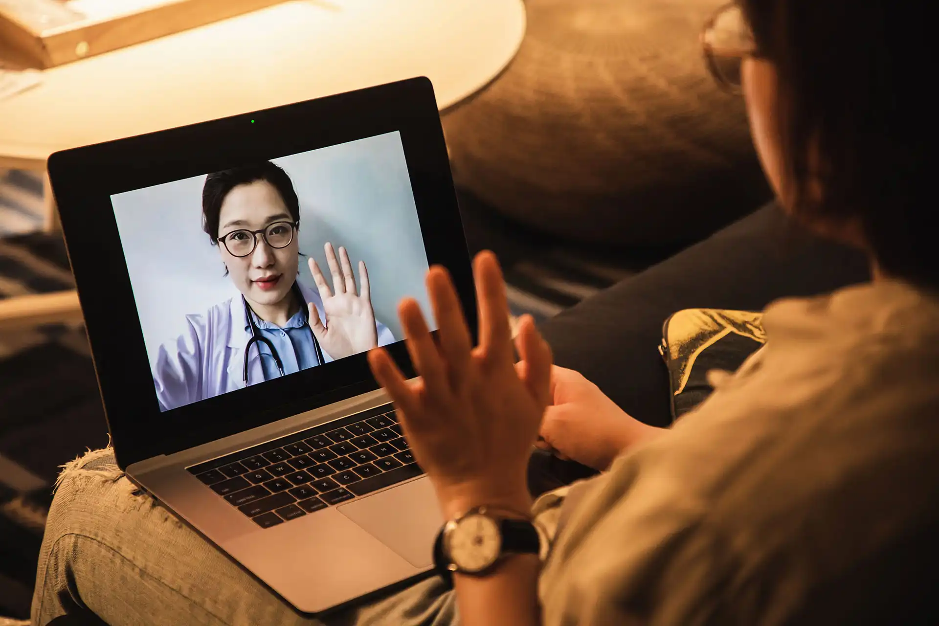 Person waving at a doctor with glasses and a stethoscope on a video call on a laptop.