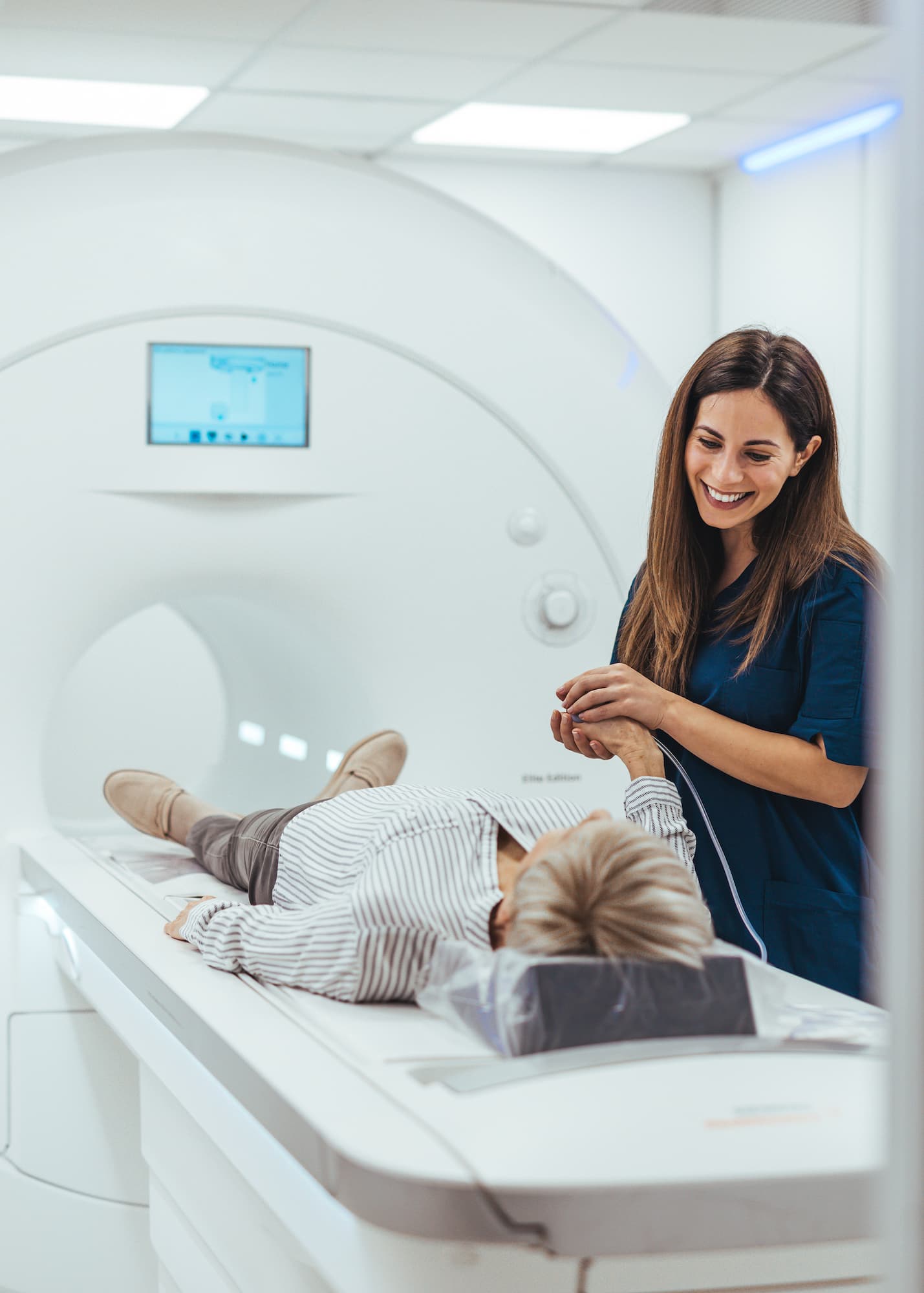Healthcare professional smiling and holding the hand of a patient lying on an MRI scanner bed.