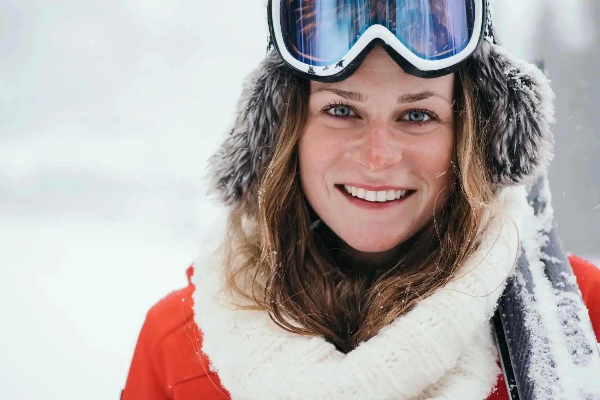 Smiling woman wearing winter gear, including a fur hat, ski goggles, and a white scarf with snow on her hair and clothes.
