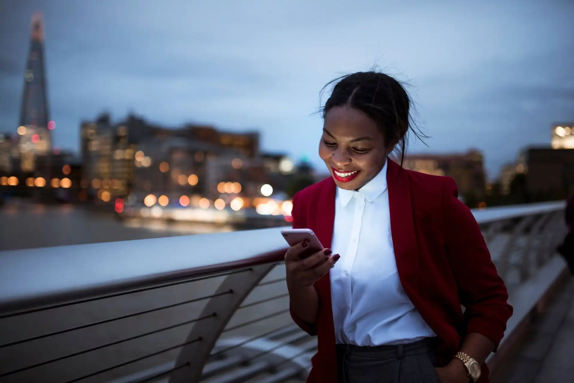 Smiling woman in a red blazer looking at her smartphone on a London city bridge at dusk.