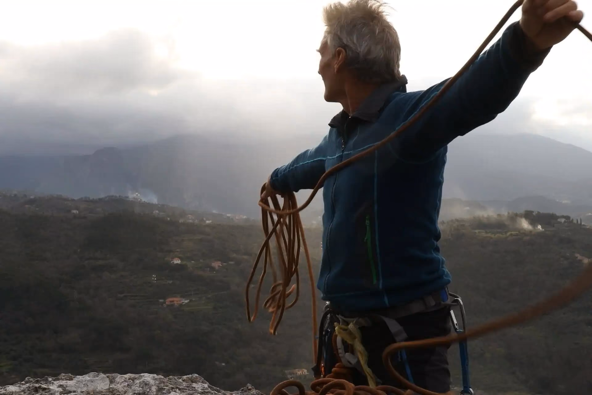 Man in climbing gear holding a rope, looking over a mountainous landscape under cloudy sky.