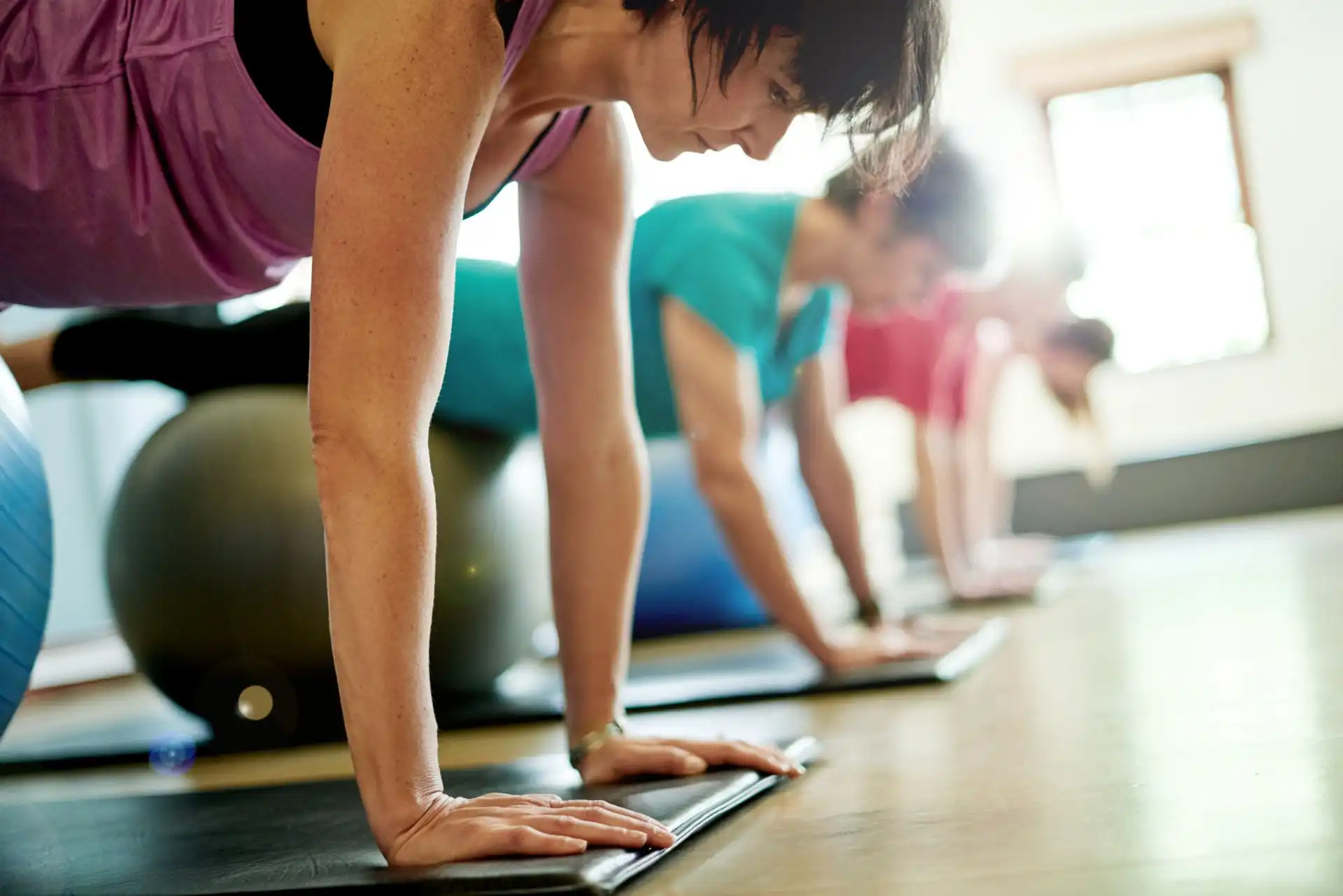 Three people doing plank exercises on mats with stability balls behind them in a bright fitness studio.