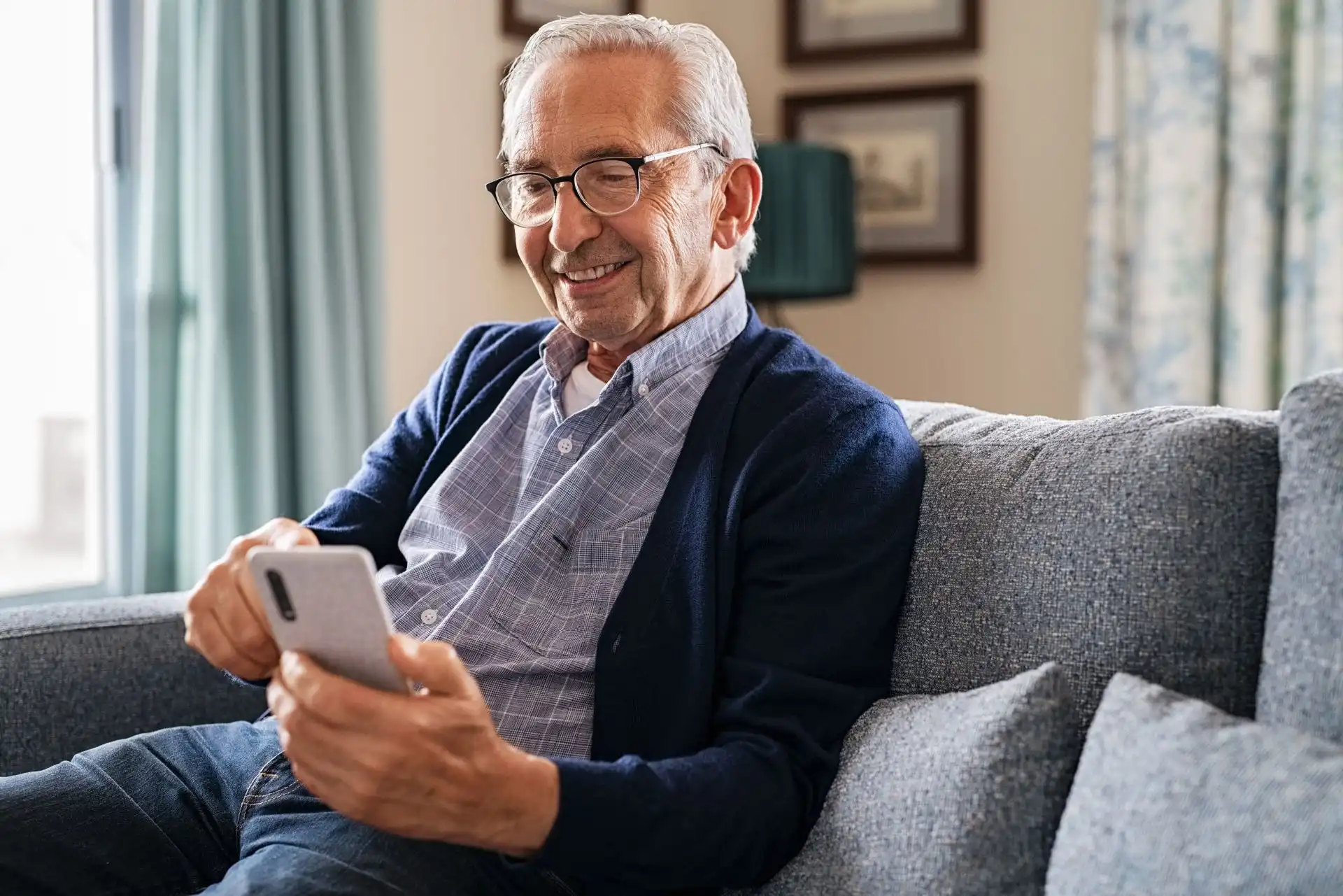 Elderly man smiling and sitting on a couch using a smartphone.