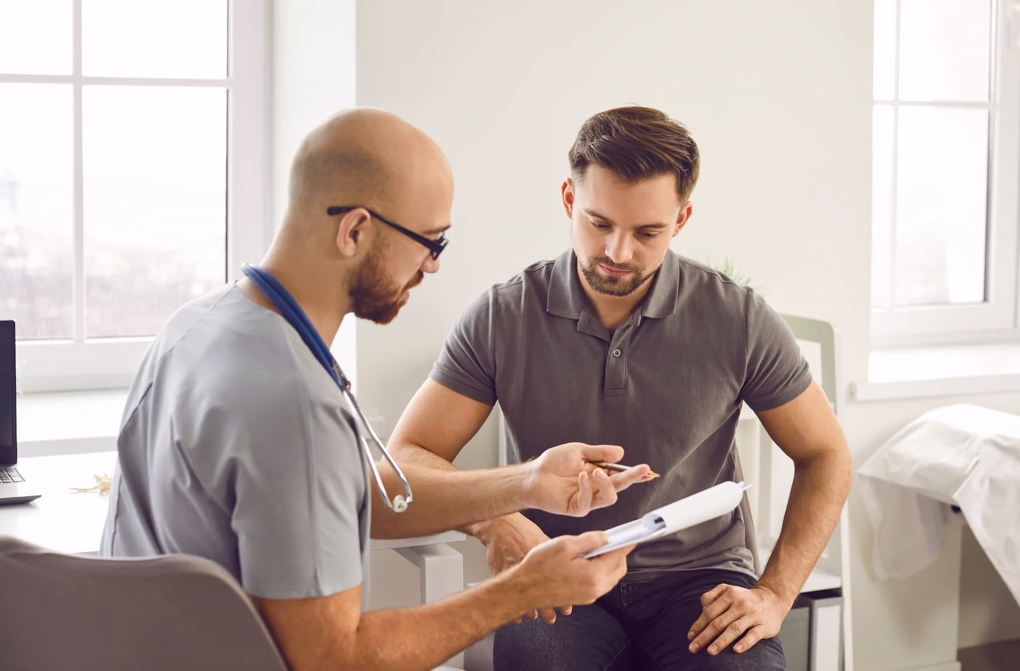 Doctor with stethoscope discussing medical information with a male patient in a bright consultation room.