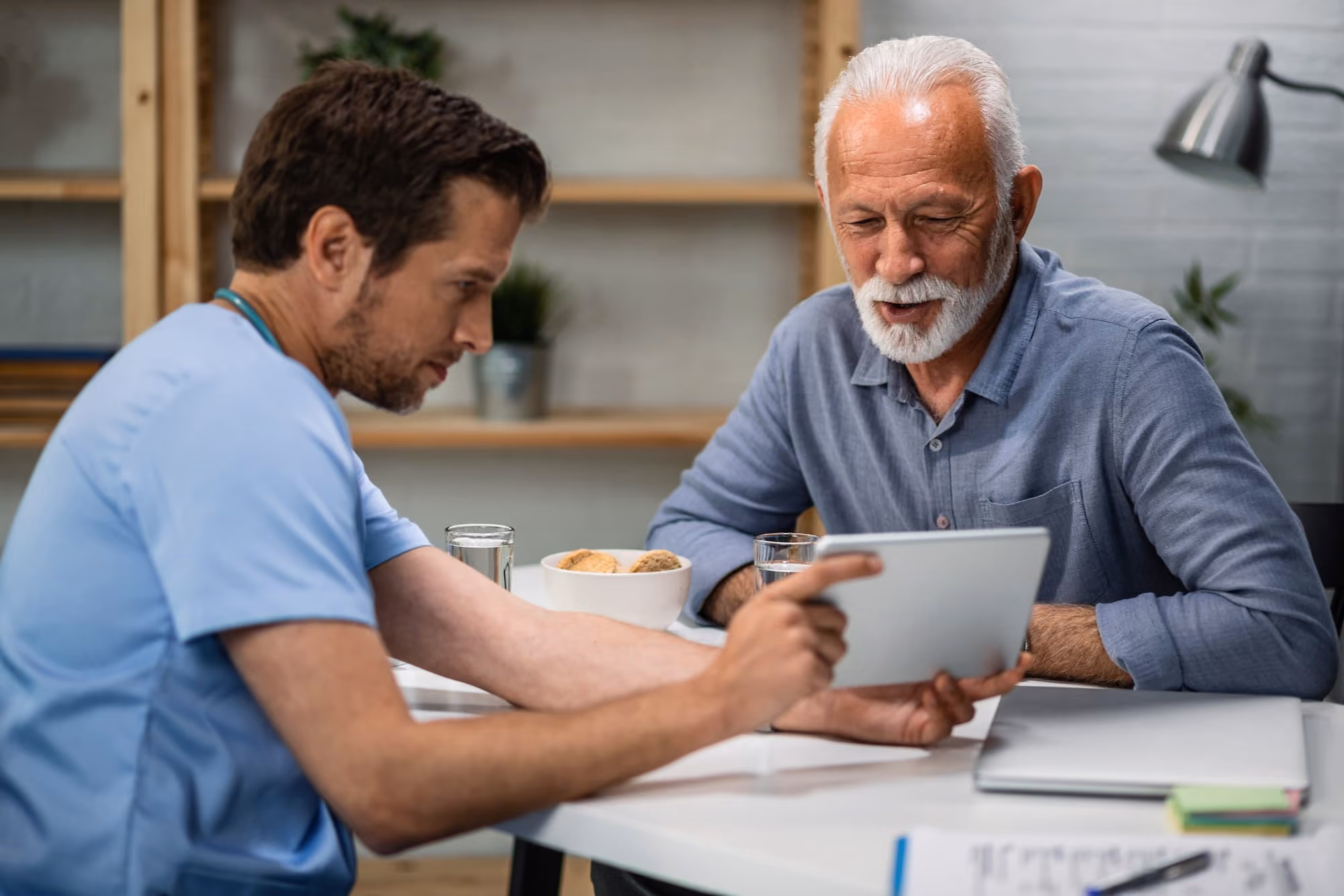 Healthcare professional showing a tablet to an elderly man at a table in a modern office.