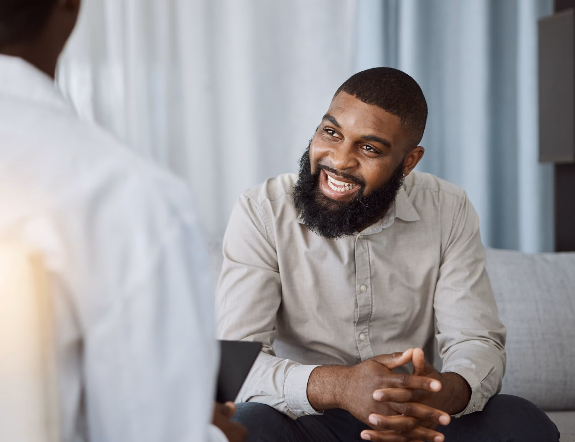 Smiling bearded man in a light shirt sitting and talking with a person in white in a cozy room.