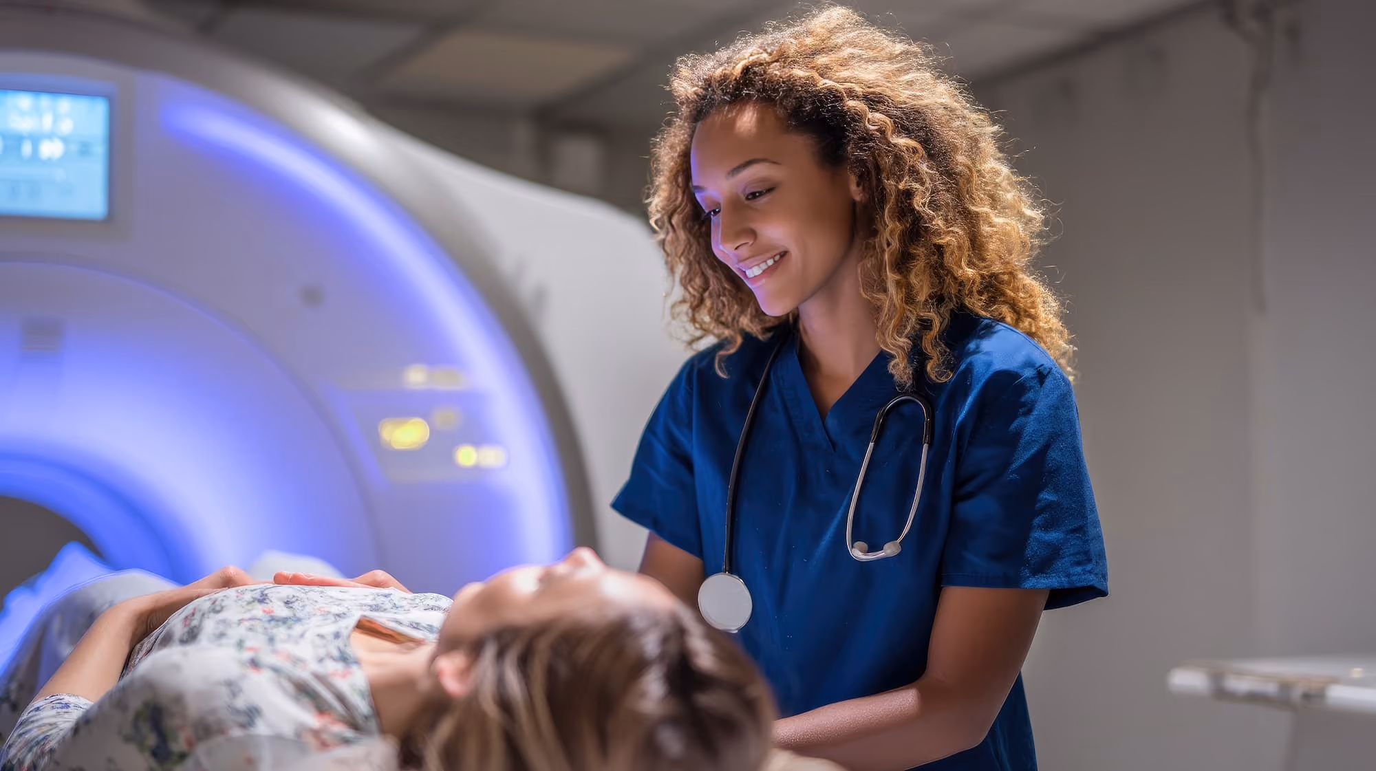 Smiling medical professional in blue scrubs with stethoscope comforting patient lying near an MRI scanner.