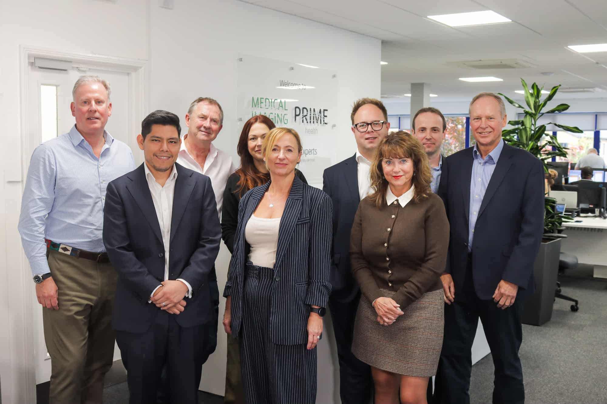 Group of nine professionally dressed Prime Health team members standing and smiling in an office with desks and computers in the background.