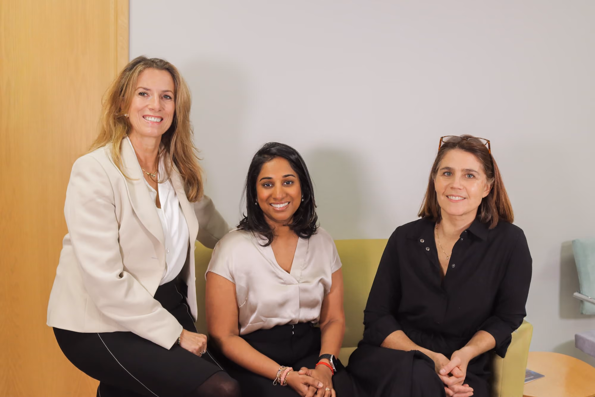 Three female Prime Health doctors smiling, two seated on a green sofa and one sitting on the armrest, dressed in business casual attire.