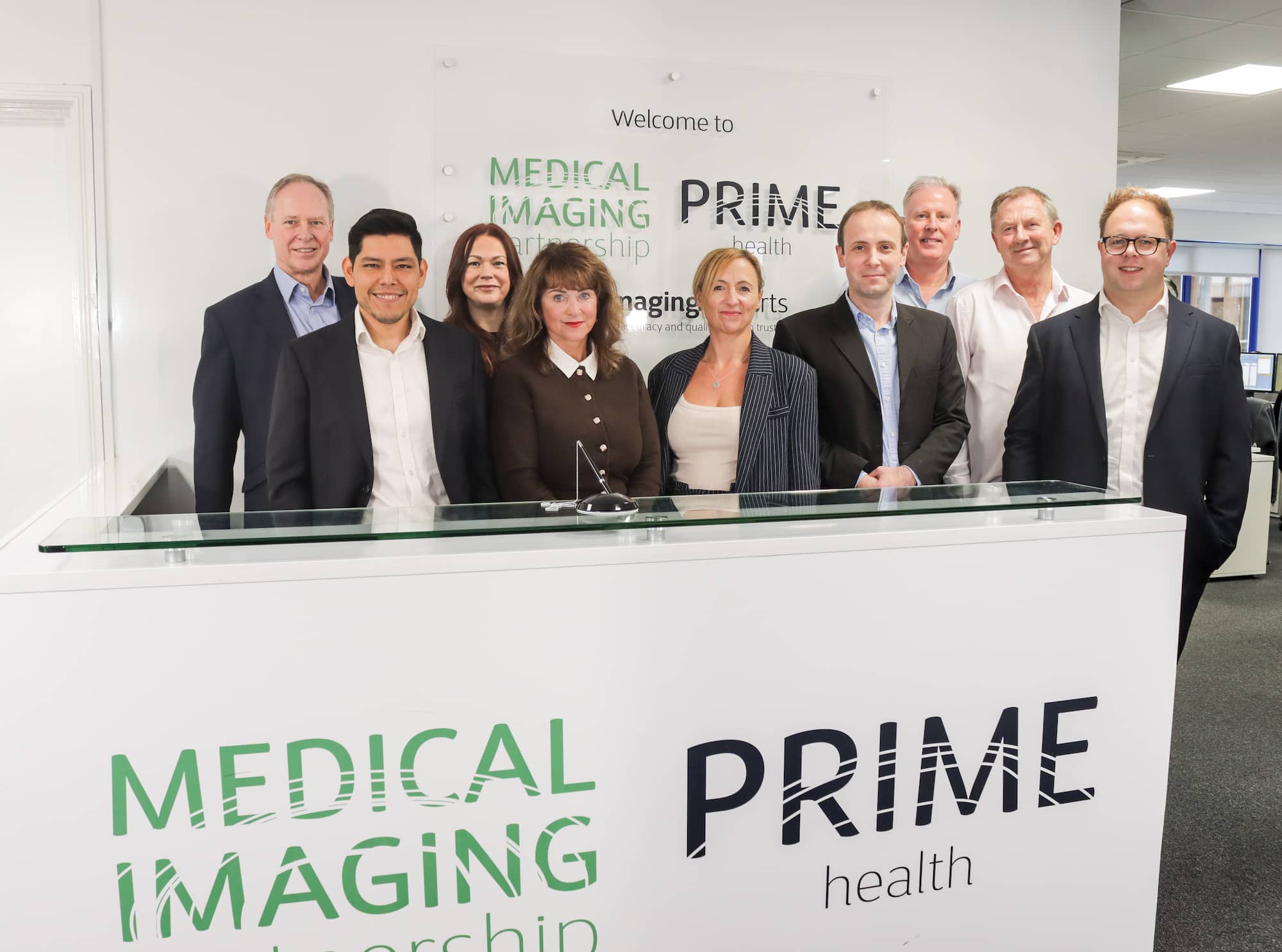 Group of nine Prime health staff members, men and women standing behind a reception desk with Medical Imaging Partnership and Prime Health logos.