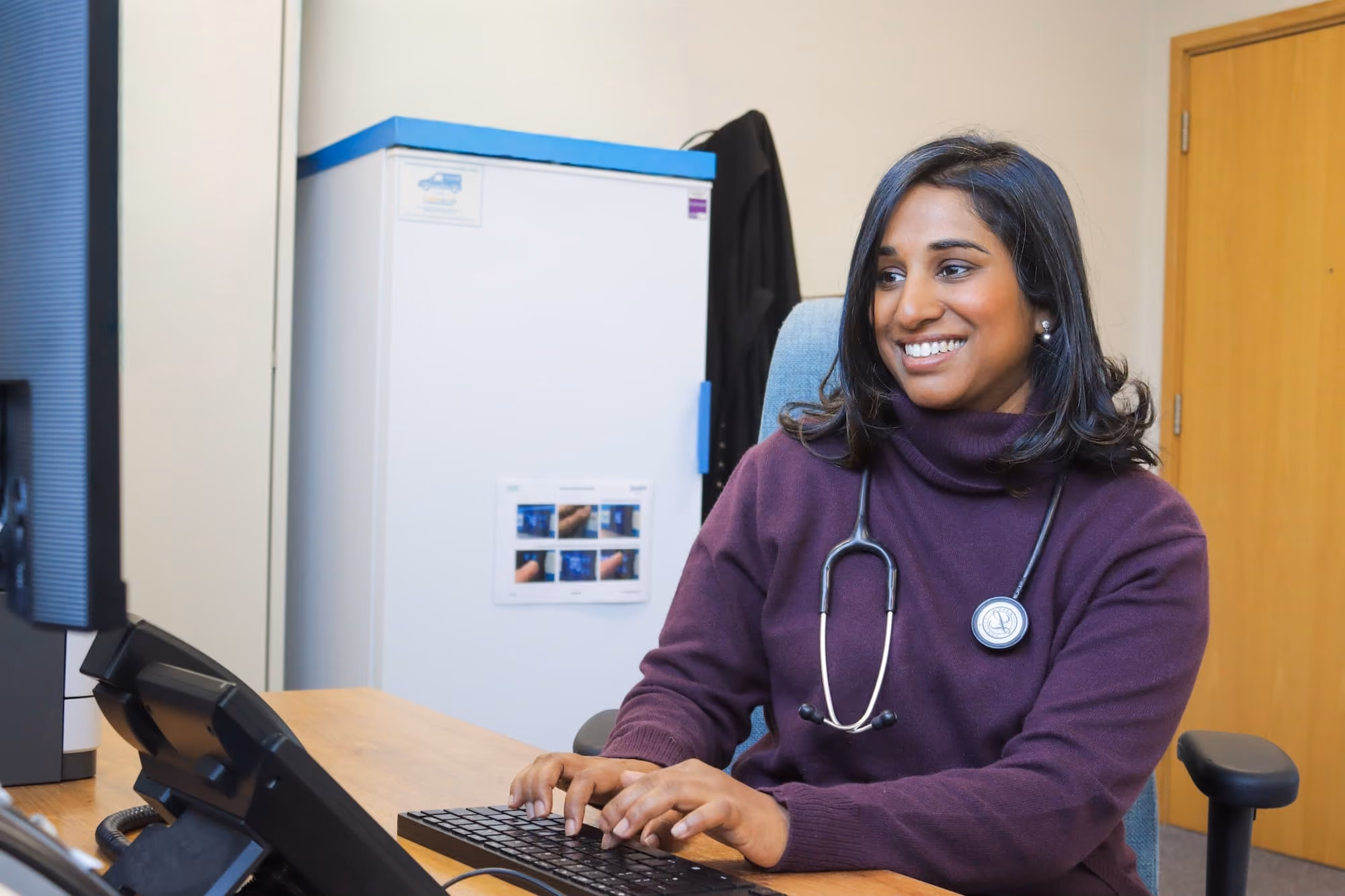 Smiling female doctor wearing a stethoscope typing on a keyboard at her desk in an office.