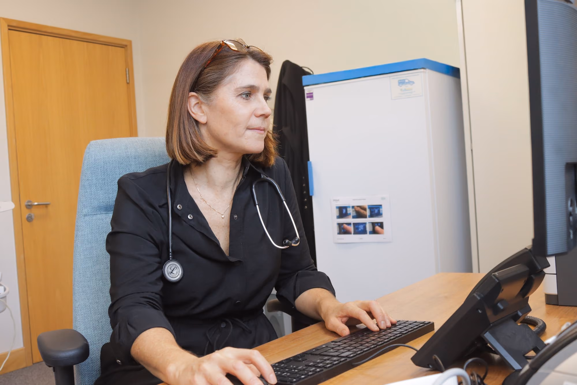 Female doctor sitting at desk, typing on a keyboard and looking at a computer screen with a stethoscope around her neck.