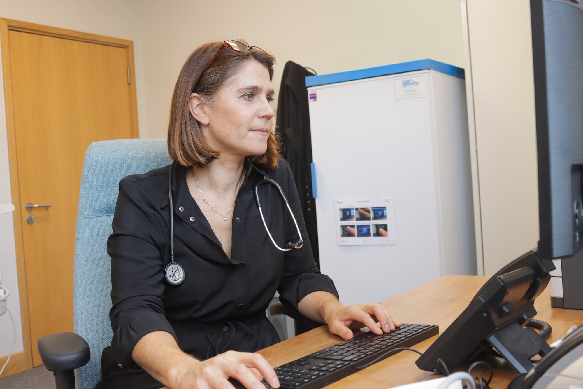 Female doctor sitting at desk, typing on a keyboard and looking at a computer screen with a stethoscope around her neck.