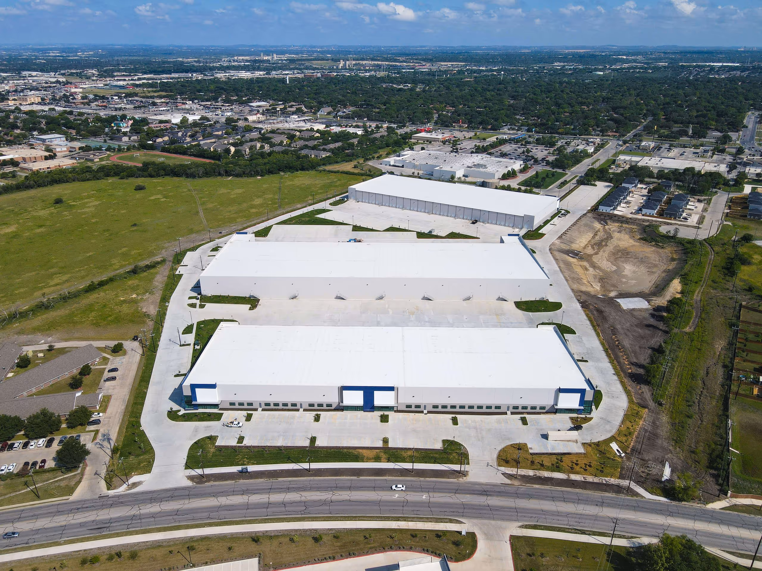 Aerial view of three large white-roofed warehouse buildings surrounded by parking lots and green fields with a cityscape in the background.