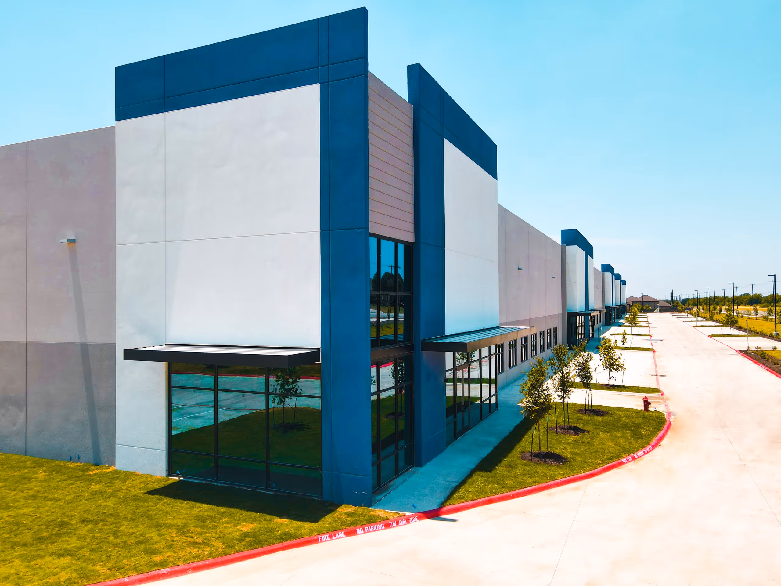 Exterior of a modern commercial building with large glass windows, blue accents, and a long driveway bordered by young trees and green grass.