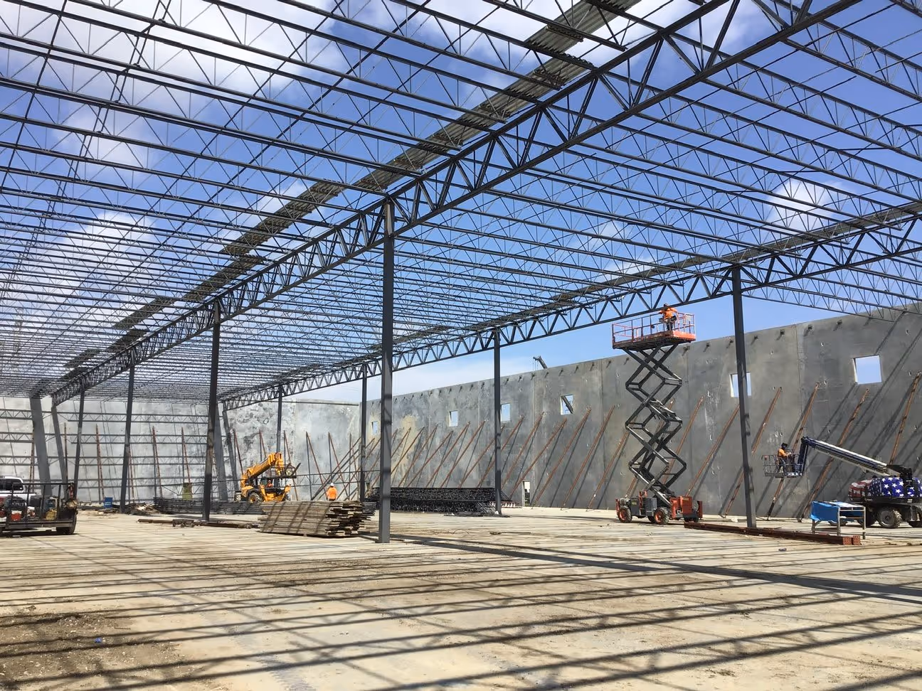 Large warehouse construction site with steel frame roof, concrete walls, and workers operating lifts.