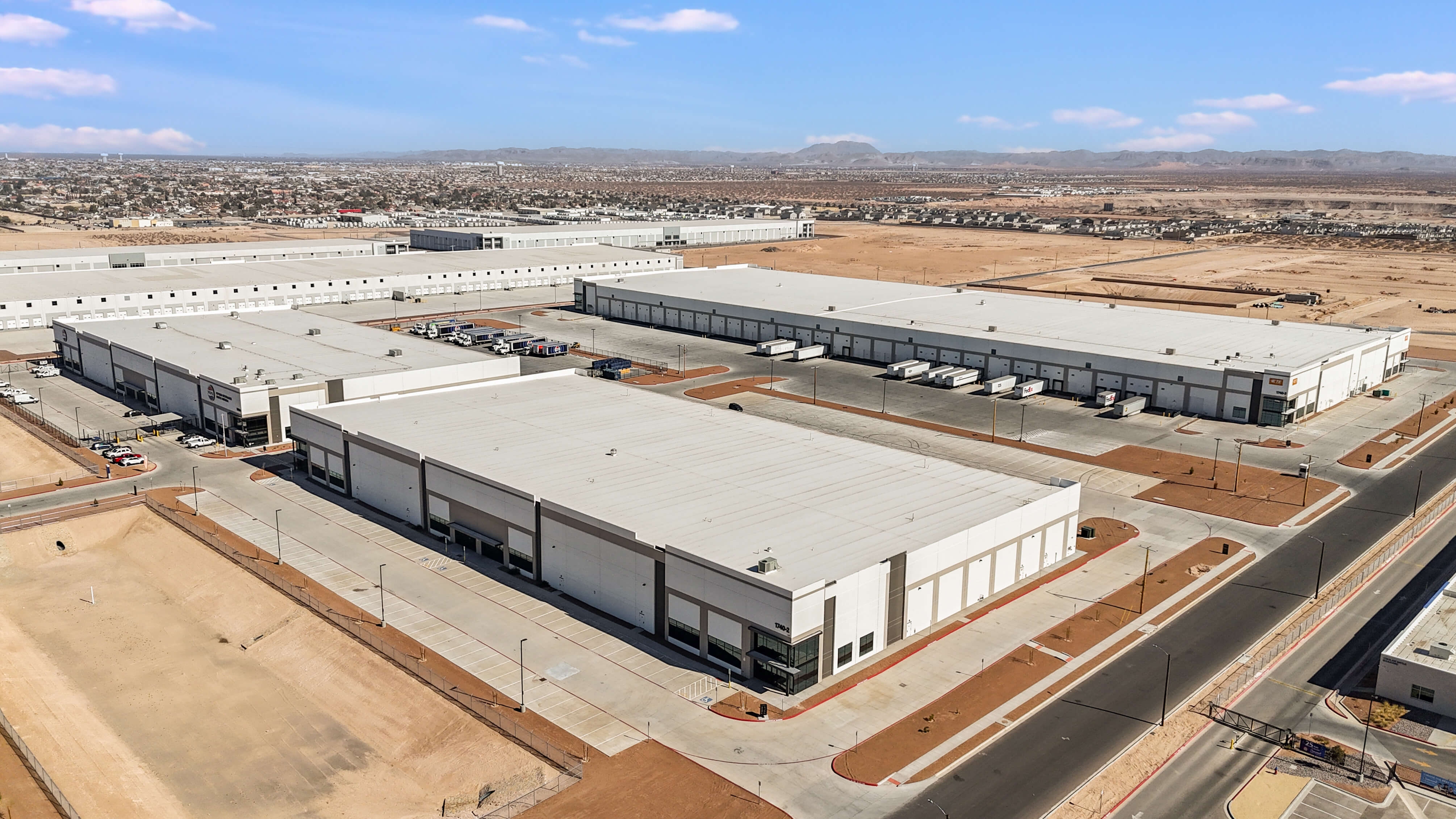Modern industrial warehouse building with blue accents, large windows, and empty parking lot under a partly cloudy sky.