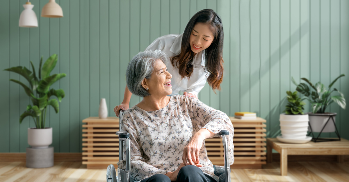 Smiling elderly woman and young girl sitting together with a smiling caregiver wearing a navy apron with 'Agensi Pekerjaan NASAV Global Sdn Bhd' text.